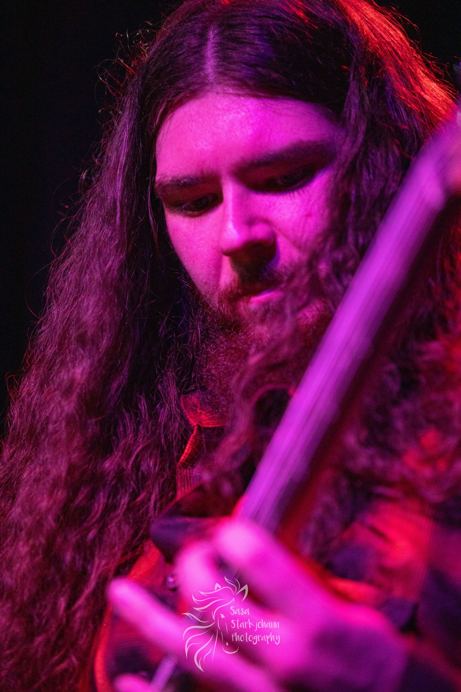 Close-up of guitarist with long hair illuminated by vibrant pink stage lighting.
