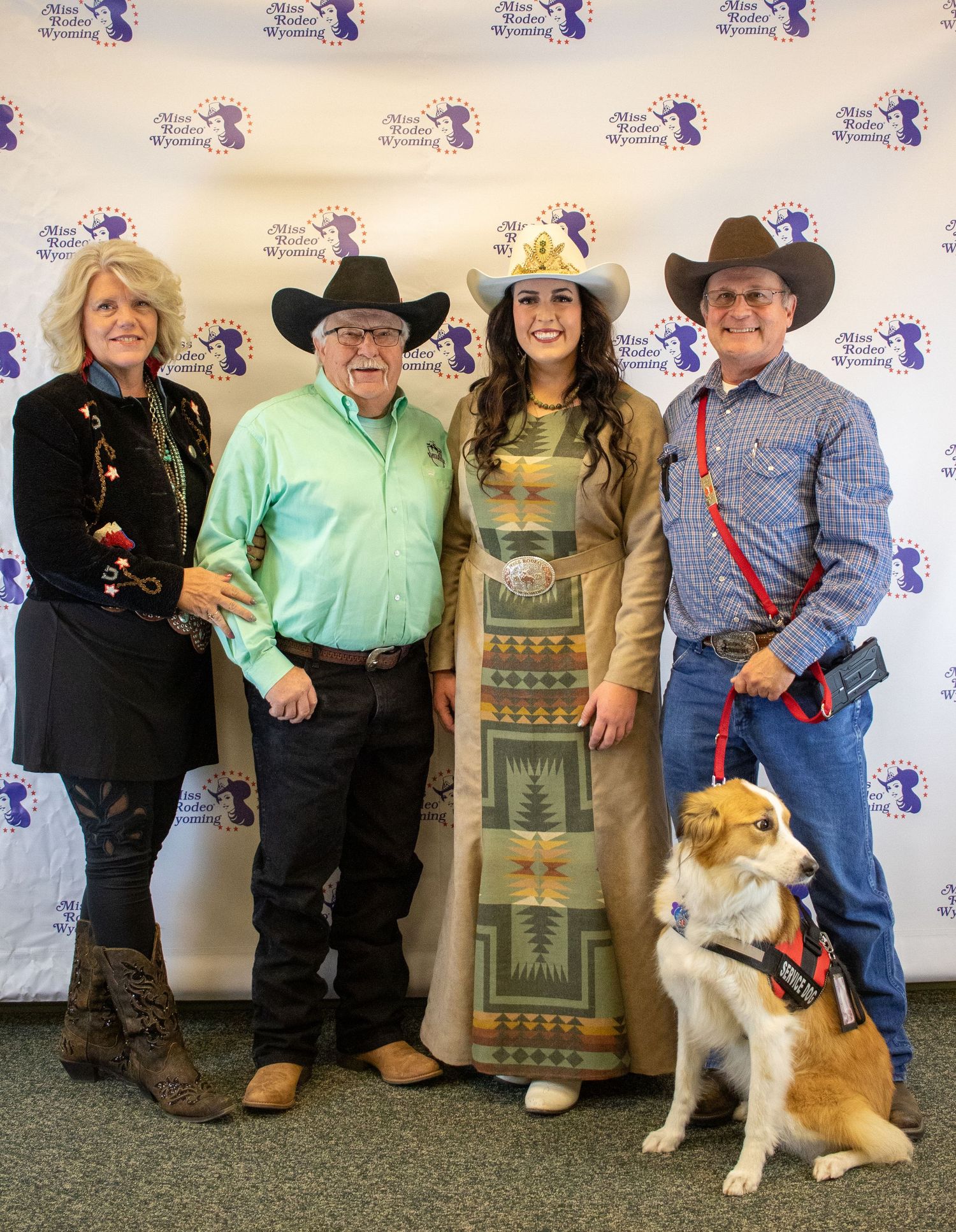 Group in western attire posing with a dog against a branded backdrop at an event.