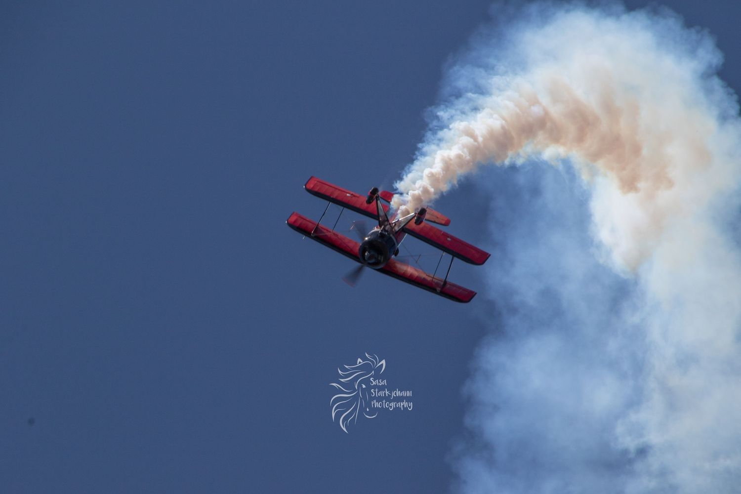 Red biplane performing aerobatic maneuver with white smoke trail against deep blue sky.