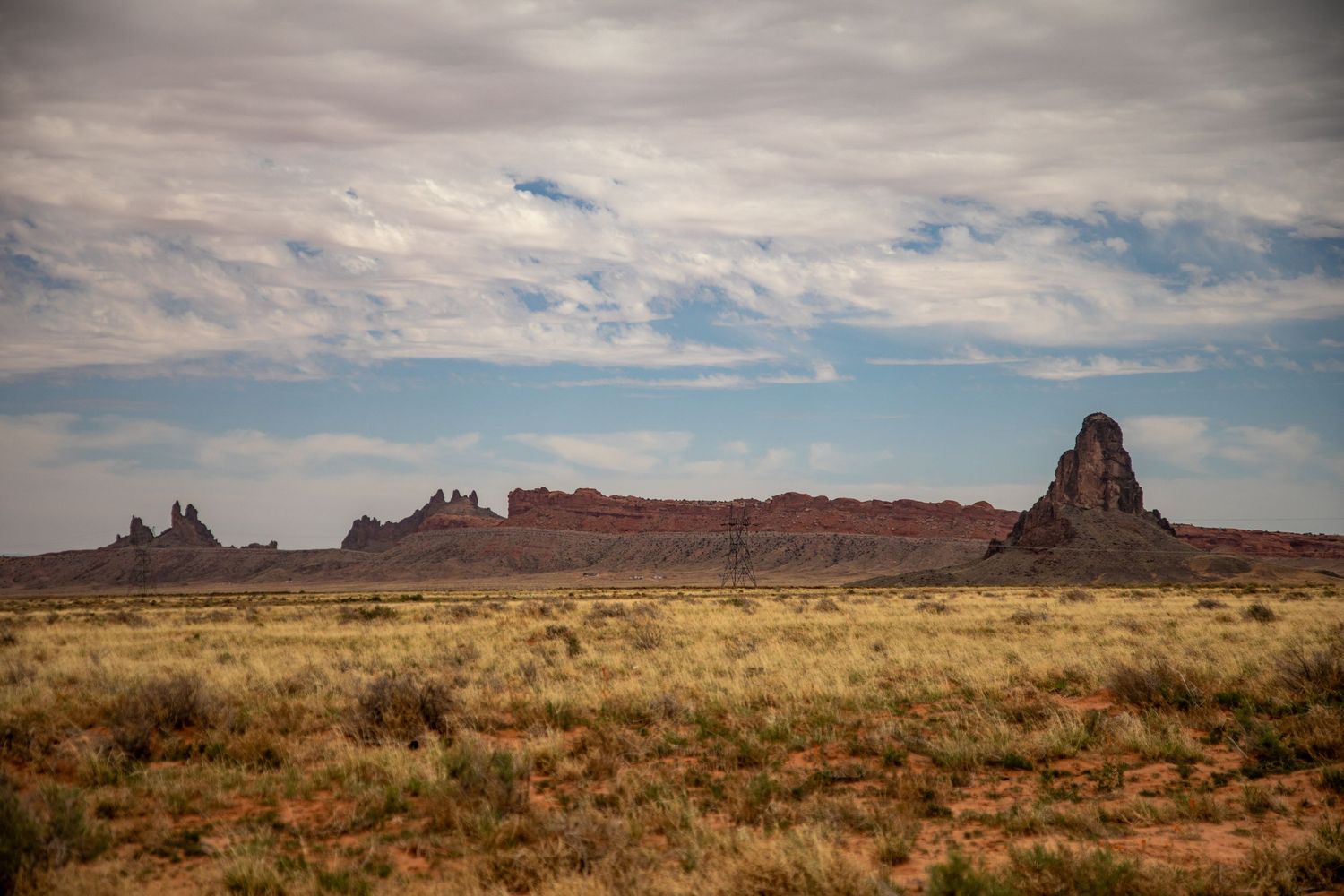Desert landscape with dramatic rock formations and mesas rising from scrubland under cloudy skies in the American Southwest.
