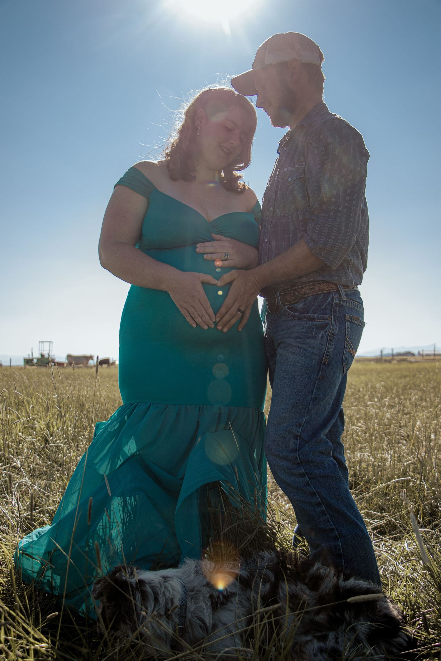 A couple stands together in a sunlit field, with the person in a flowing teal dress creating a romantic silhouette.