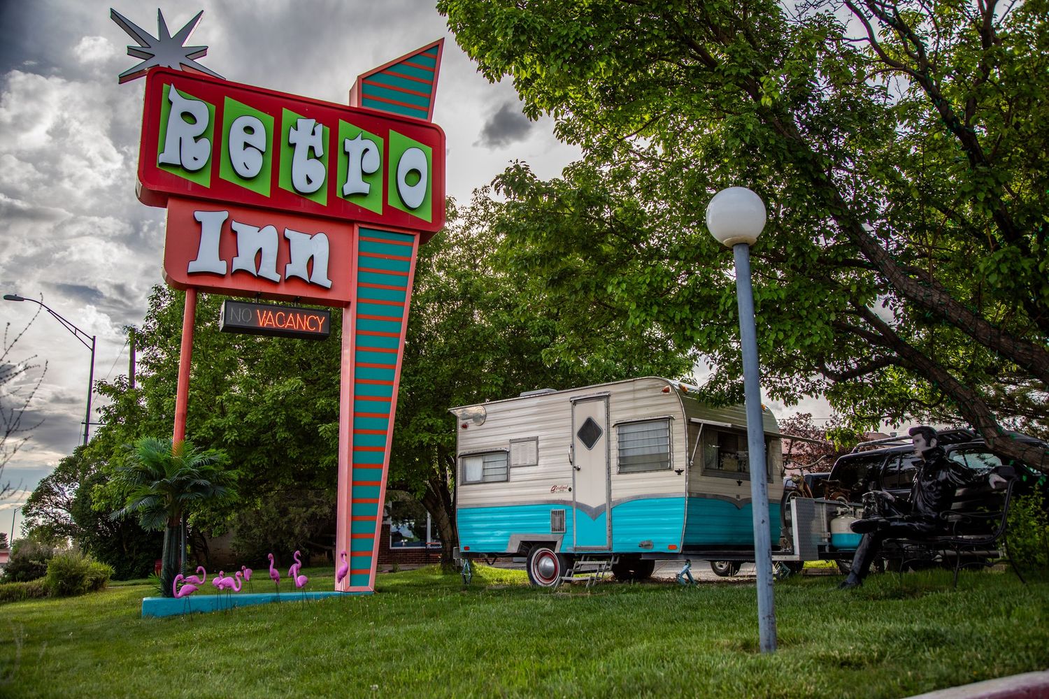 Vintage retro motel sign stands tall against cloudy sky with classic turquoise trailer parked beneath it.