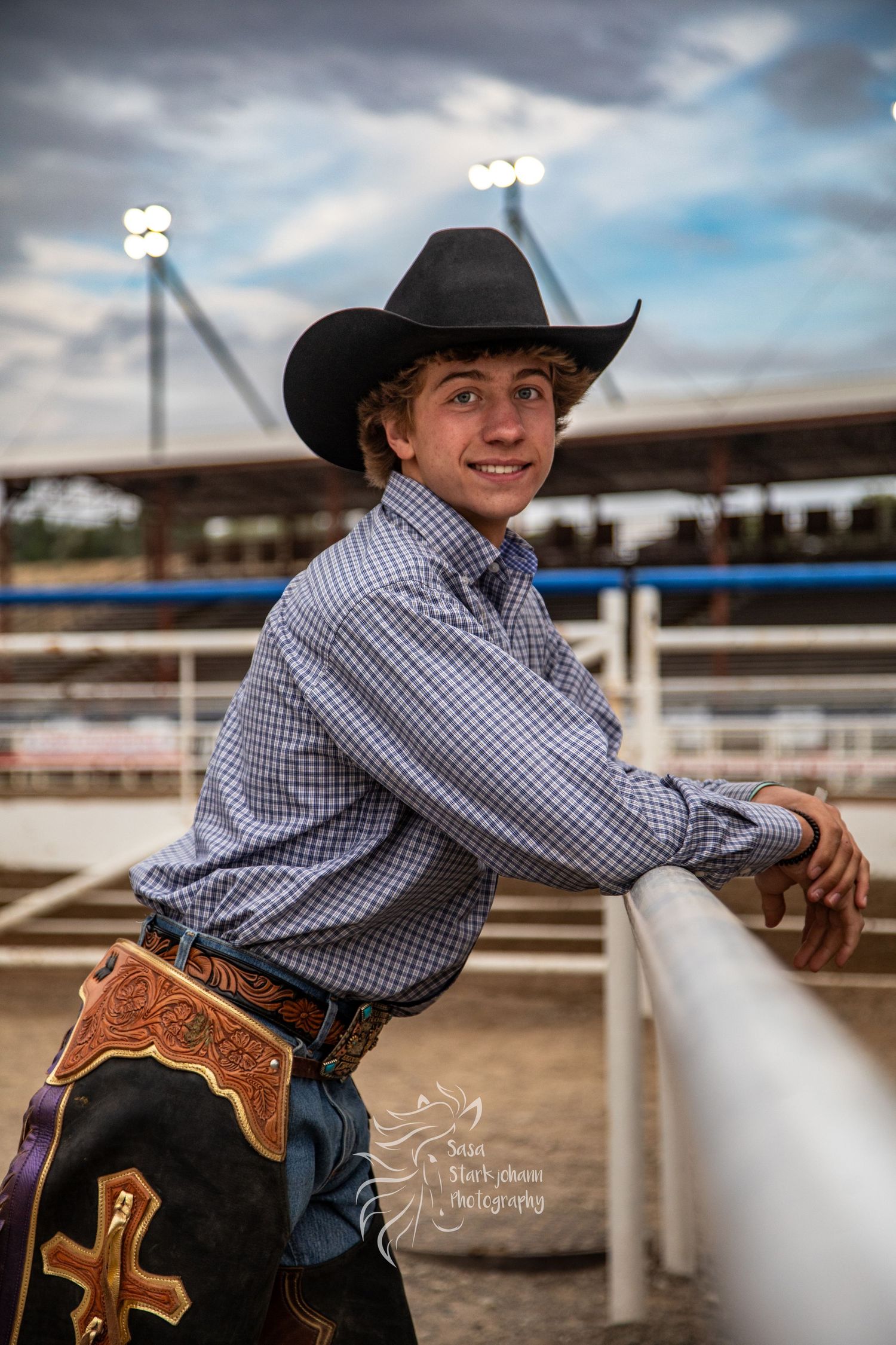 Young person in western attire and cowboy hat leans on white fence at rodeo arena during cloudy day.