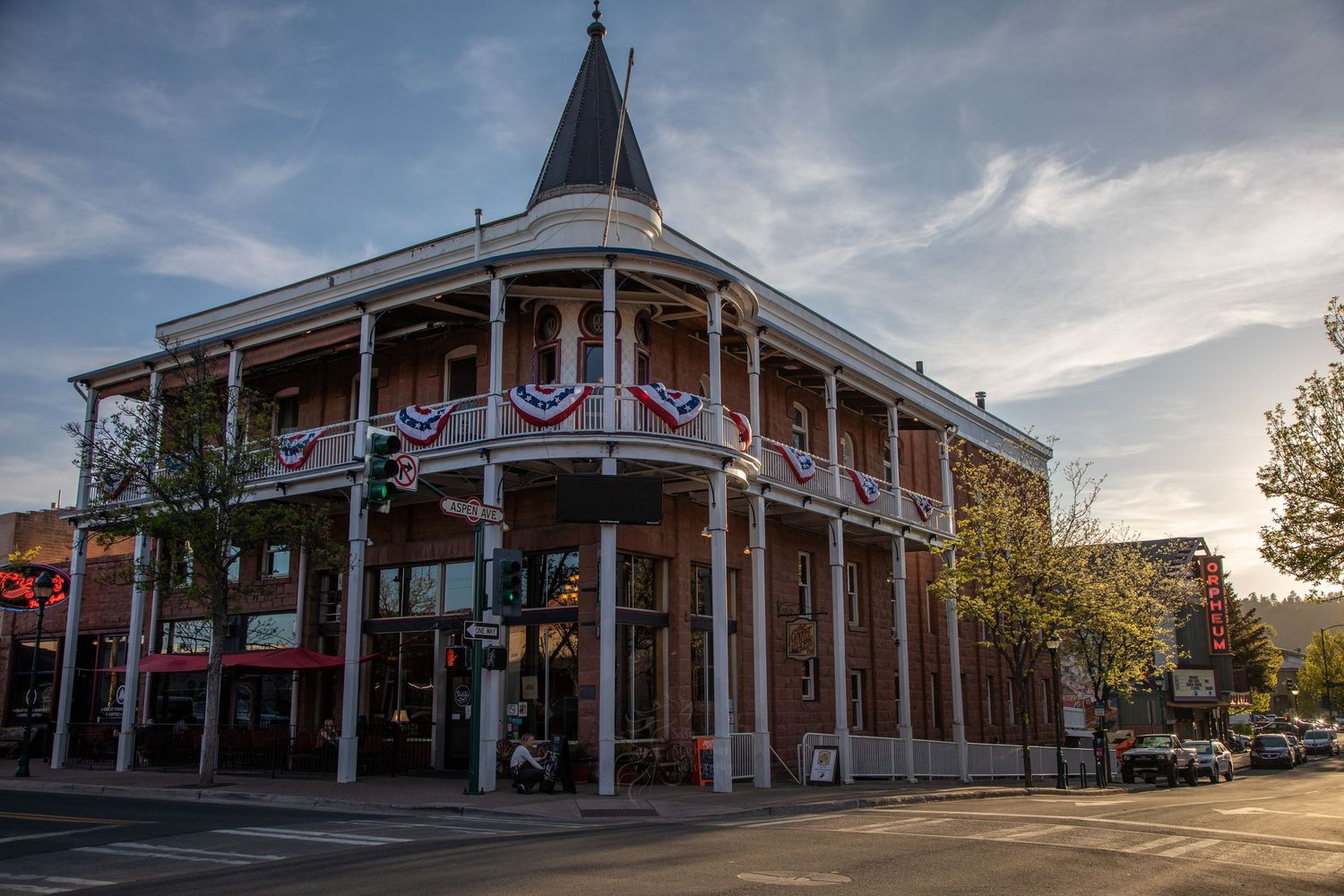 Historic two-story Victorian building with wraparound porch decorated in patriotic bunting stands on corner at sunset.