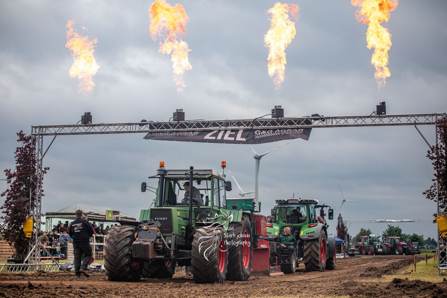 Green tractors compete in a tractor pulling event under a ZIEL finish arch with dramatic fire flames at an outdoor competition.