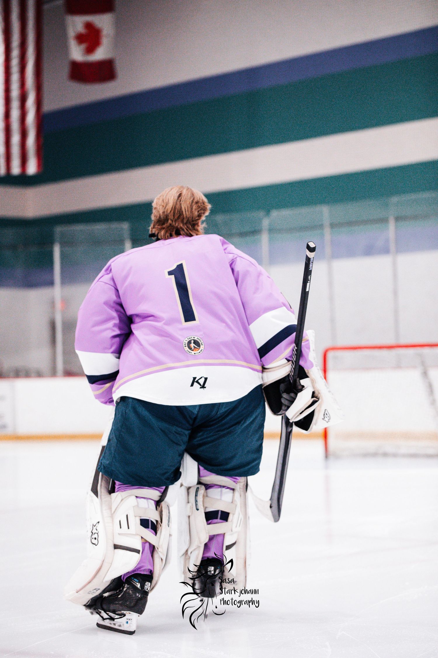 Hockey goalie in purple jersey standing on ice rink with American and Canadian flags visible.