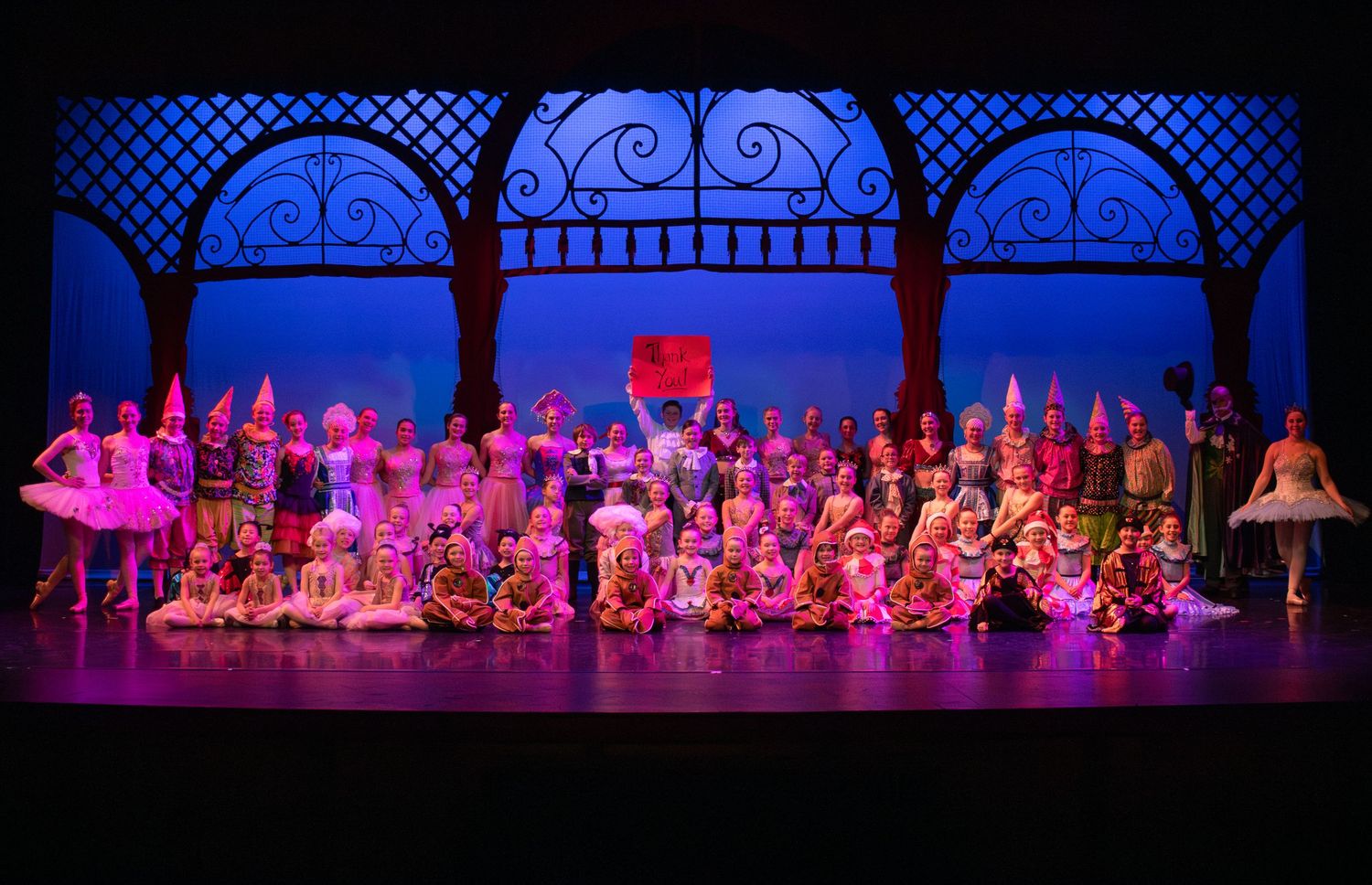 Large ensemble cast performs ballet scene on ornate stage with blue backdrop.
