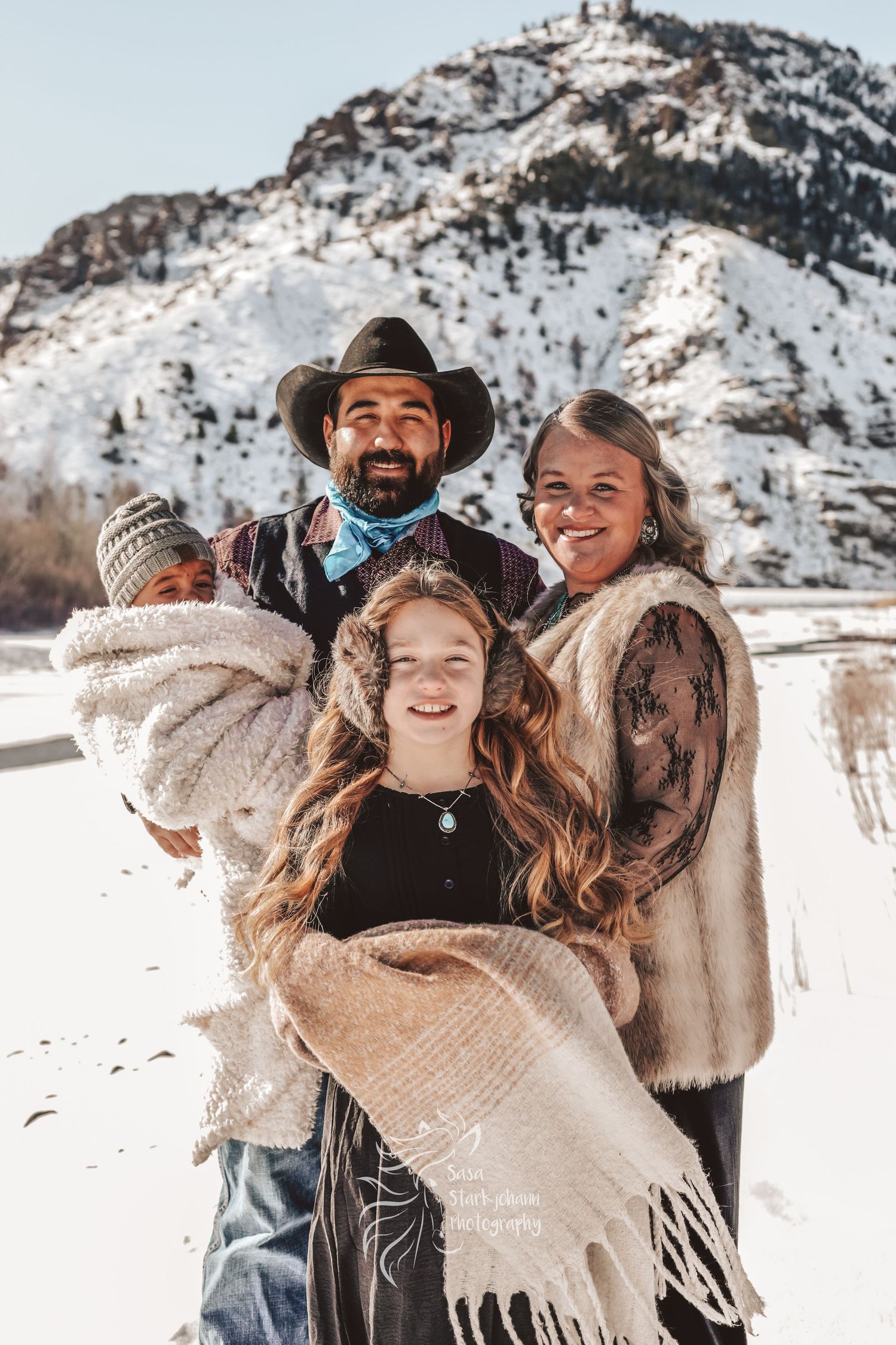 A family poses together in warm winter clothes against a snowy mountain landscape.