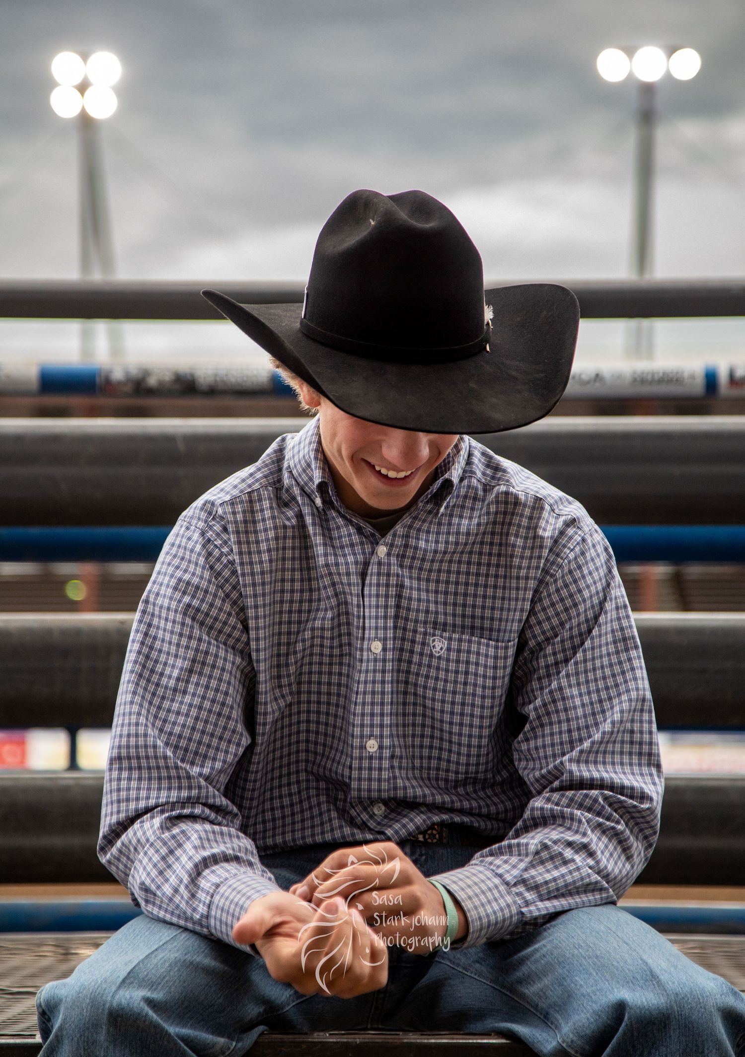 A person wearing a black cowboy hat and plaid shirt sits on bleachers while looking down at their hands.