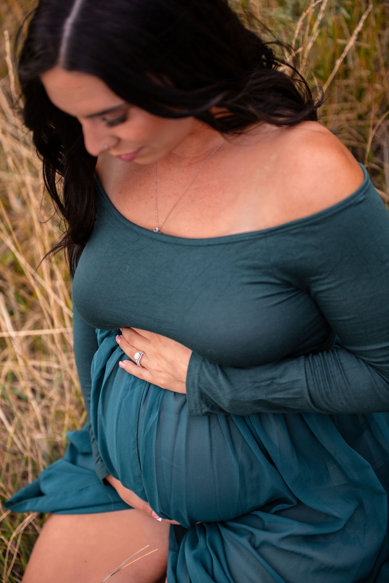 A person in a teal dress cradles their pregnant belly while standing in a field of golden grass at sunset.