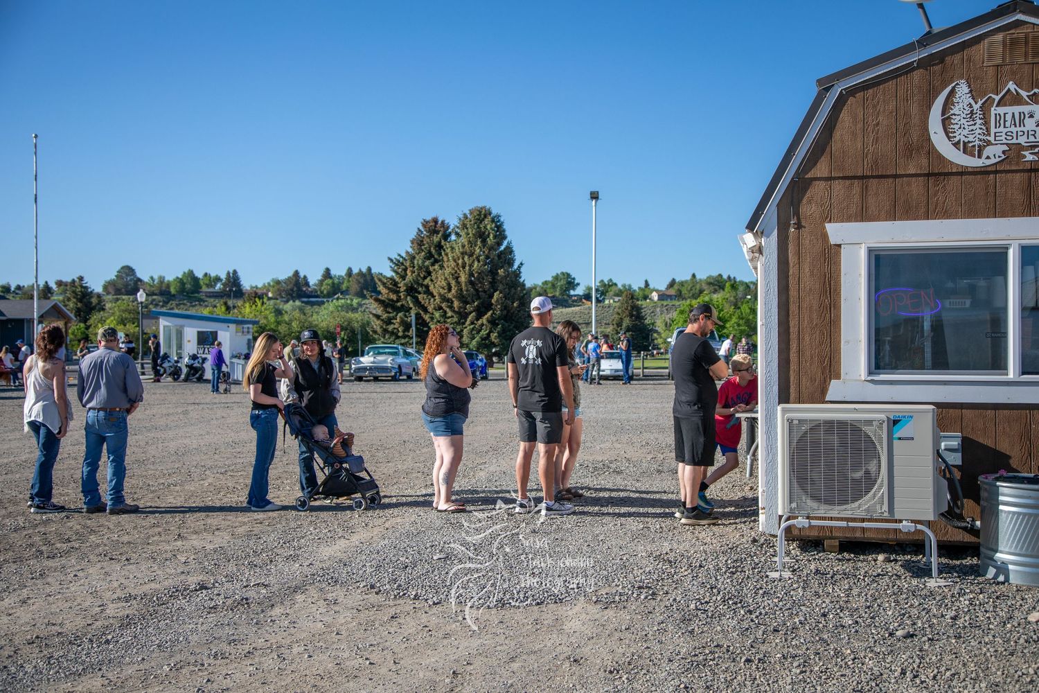 Rustic wooden farm buildings surrounded by people gathered for an outdoor event on a sunny day.