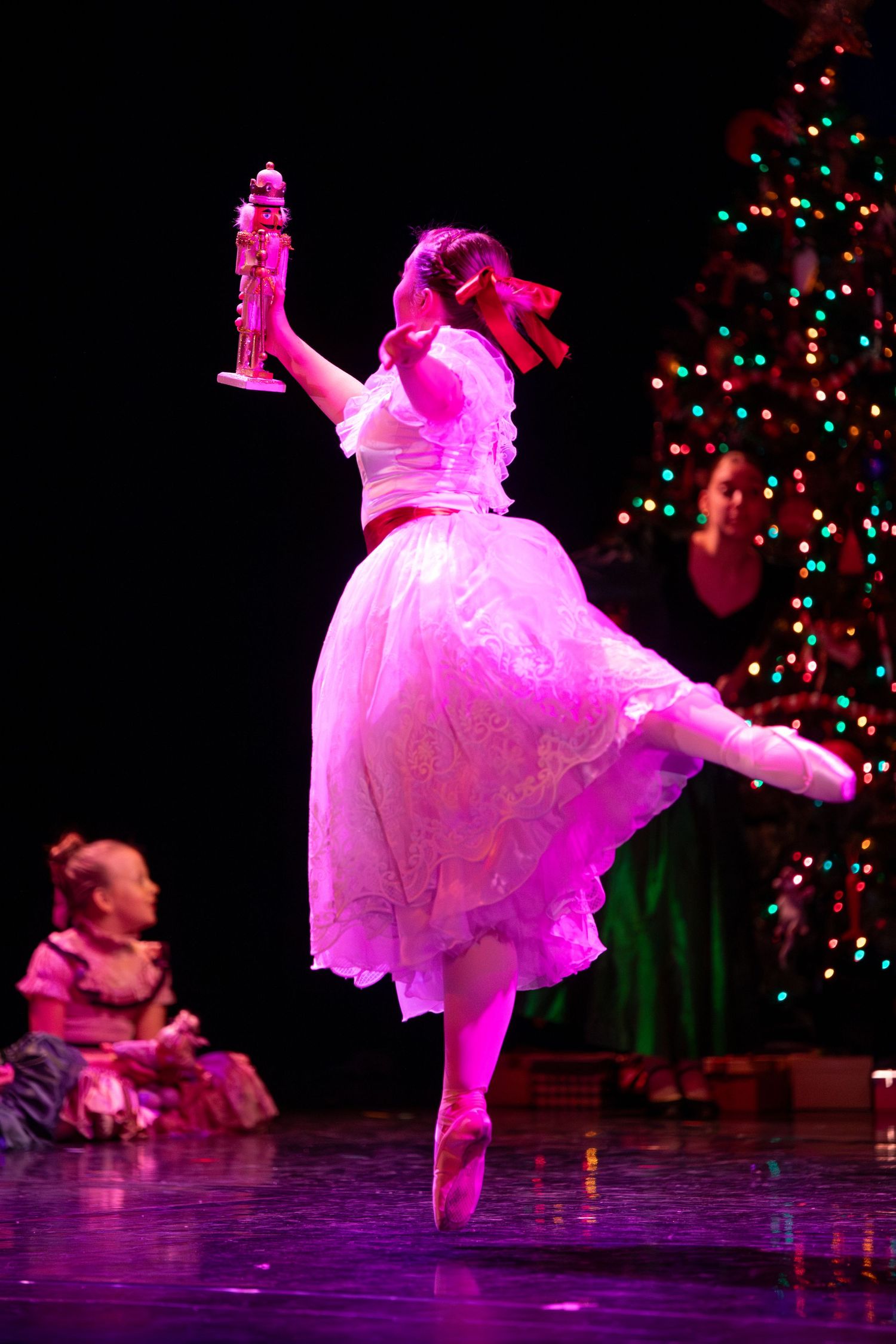 Dancer in dress performs ballet move holding prop near decorated Christmas tree.