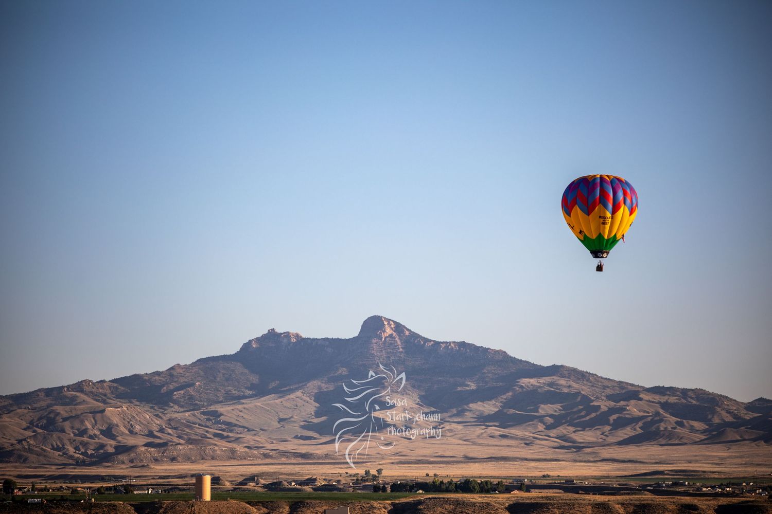 Colorful hot air balloons float above a desert mountain landscape against a clear blue sky at sunrise.