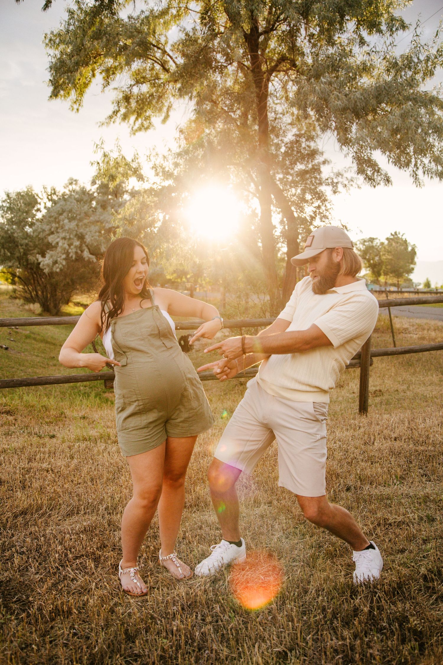 Playful couple dancing together in a park at sunset with sun flare and trees in background.
