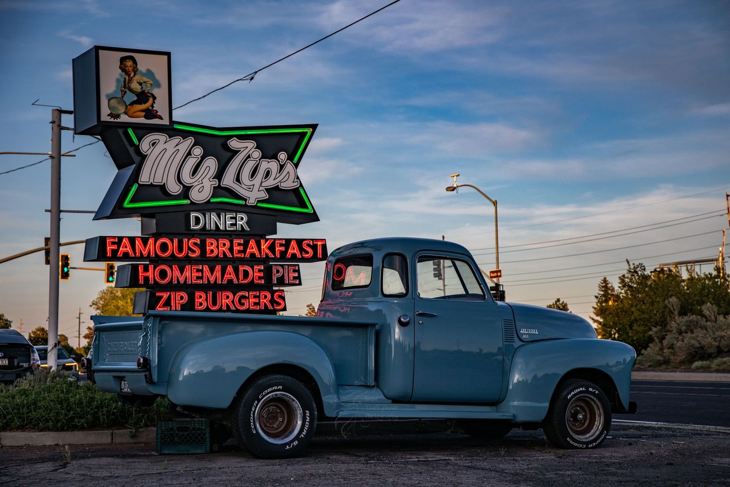 A vintage blue pickup truck parked beneath a retro diner sign advertising burgers and homemade pie against a cloudy sky.