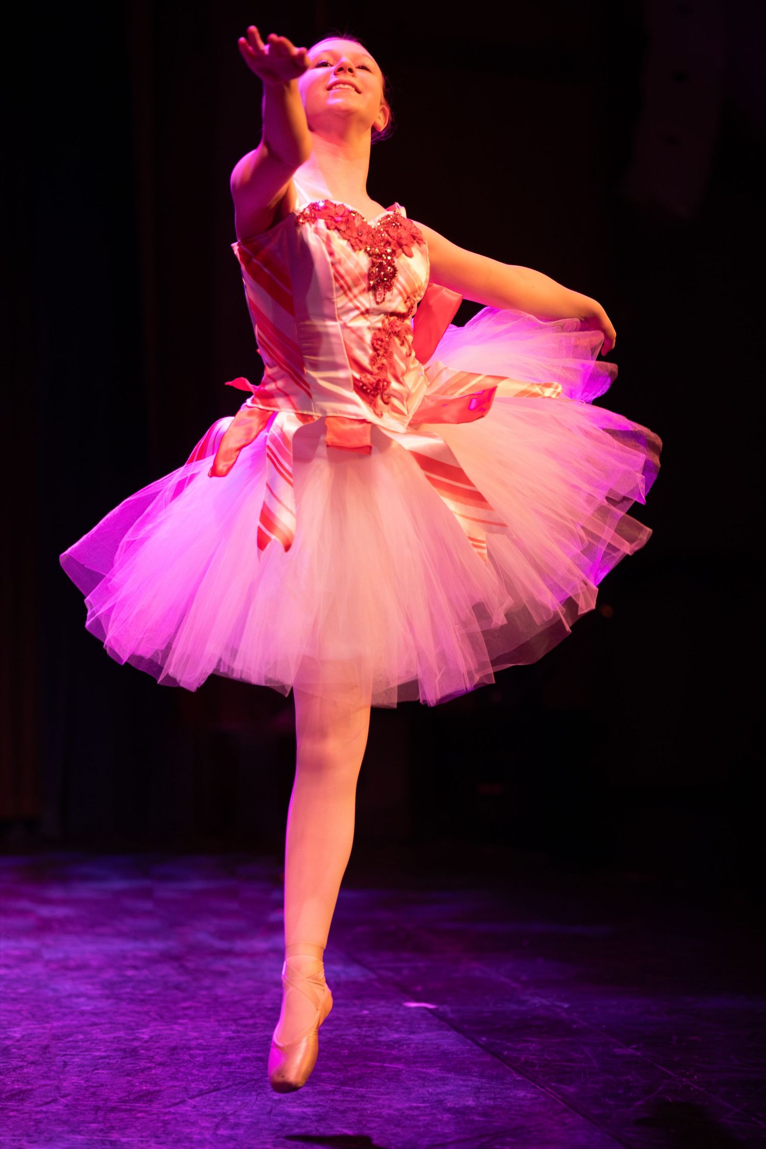 Ballet dancer in pink tutu performs graceful leap during stage performance under purple lighting.