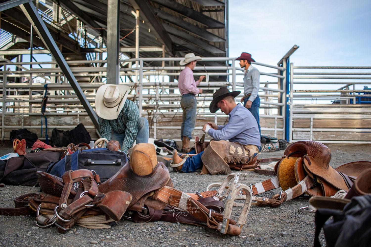 Rodeo equipment and leather gear scattered on ground near arena bleachers during western event.