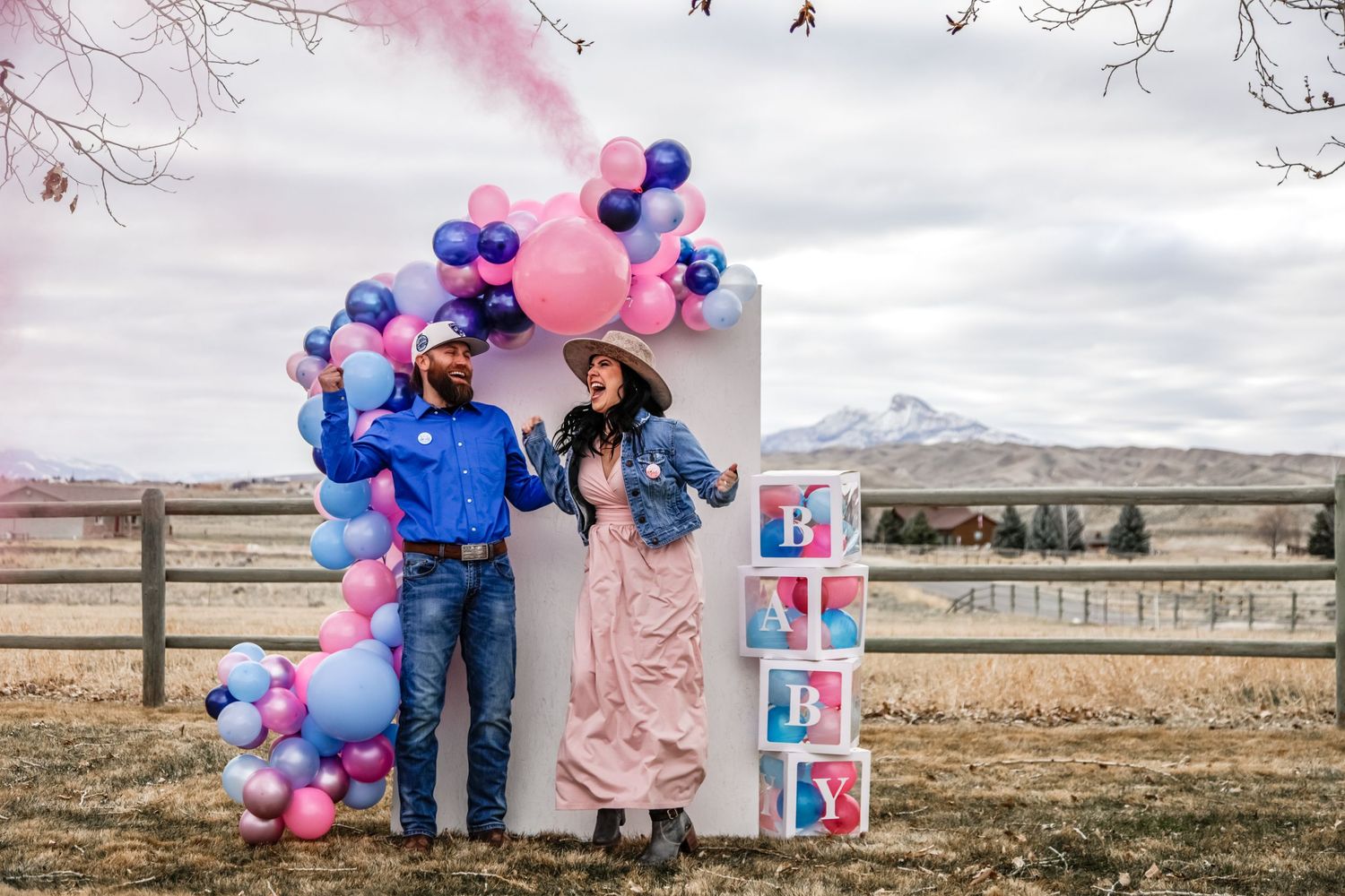 Gender reveal party setup with pink and blue balloon arch and photo display in rural setting.