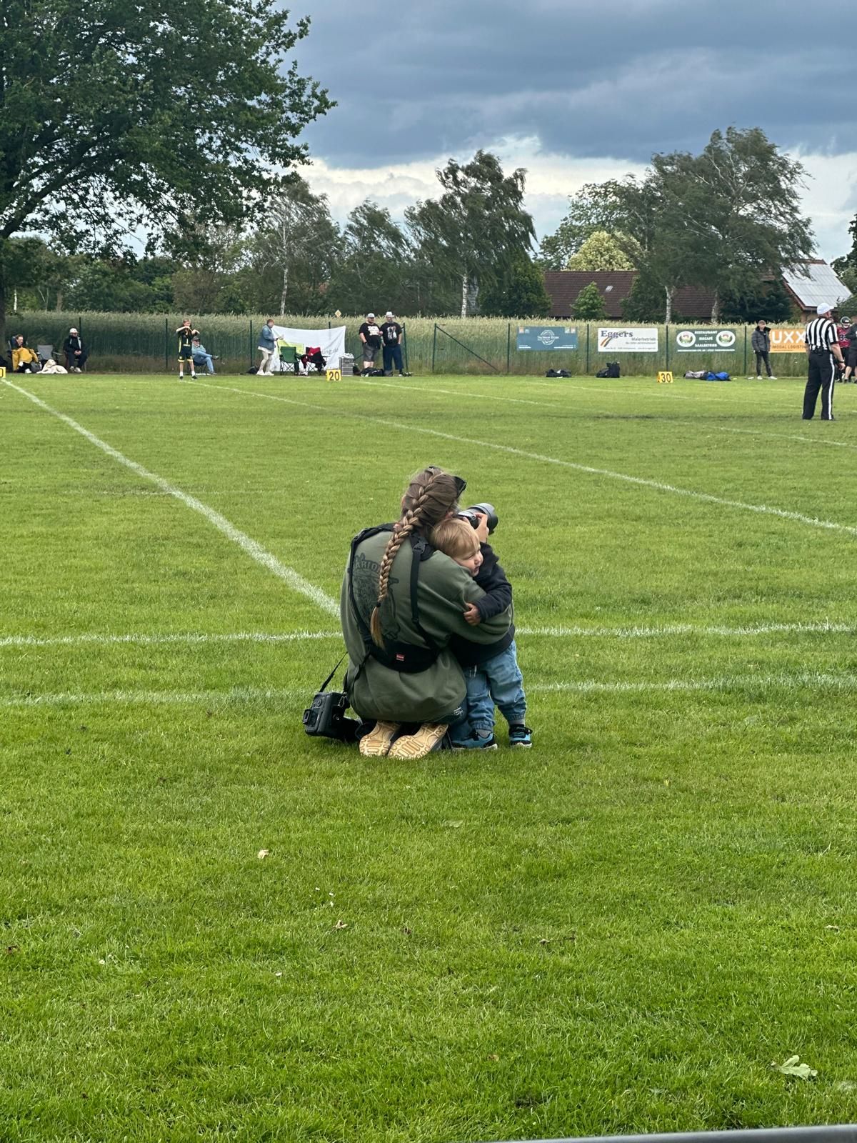 Photographer kneeling on green football field hugging a toddler during an outdoor game on a cloudy day.