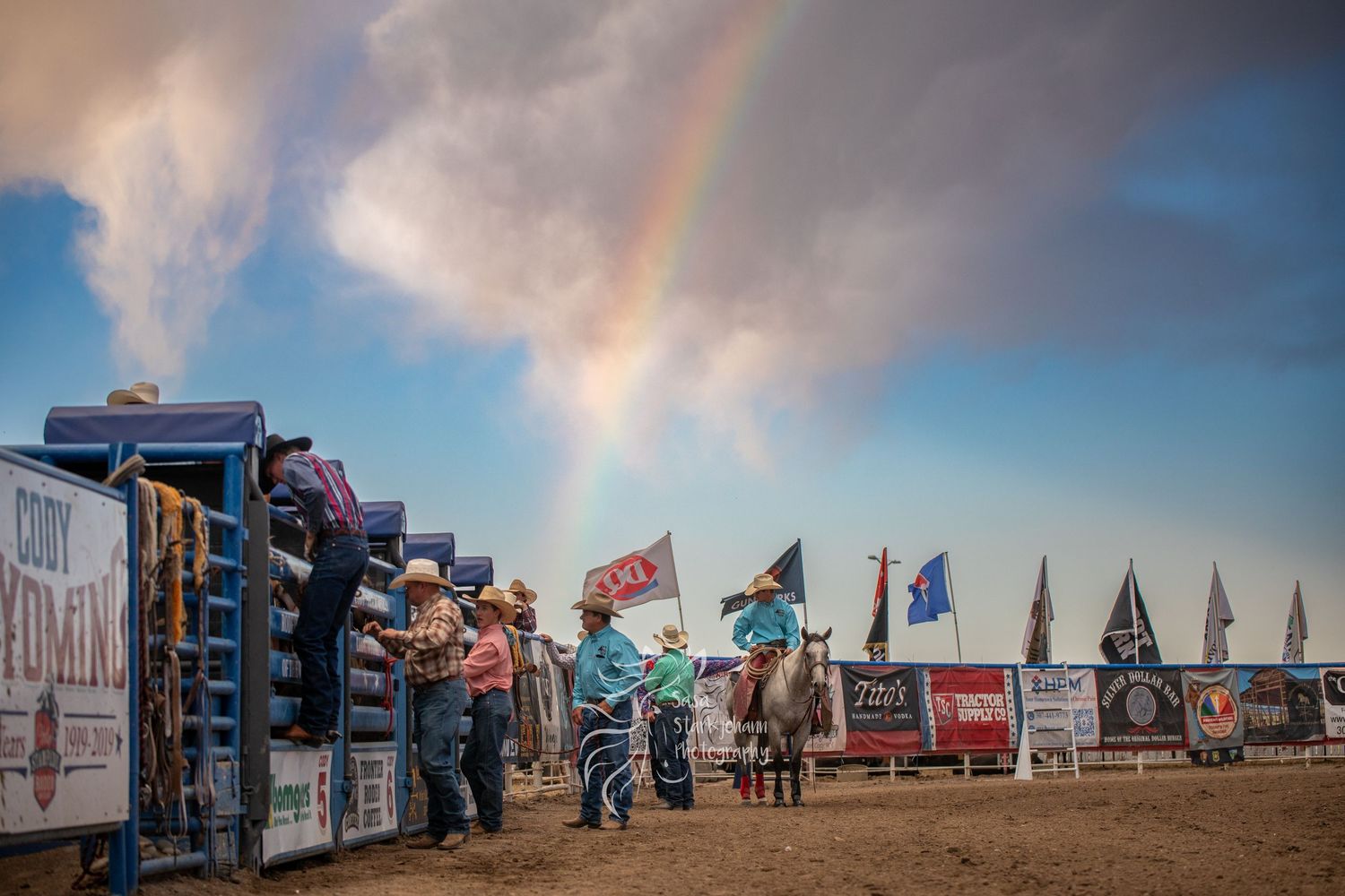 Cowboys prepare at a rodeo chute under a dramatic rainbow sky with colorful flags waving in the background.