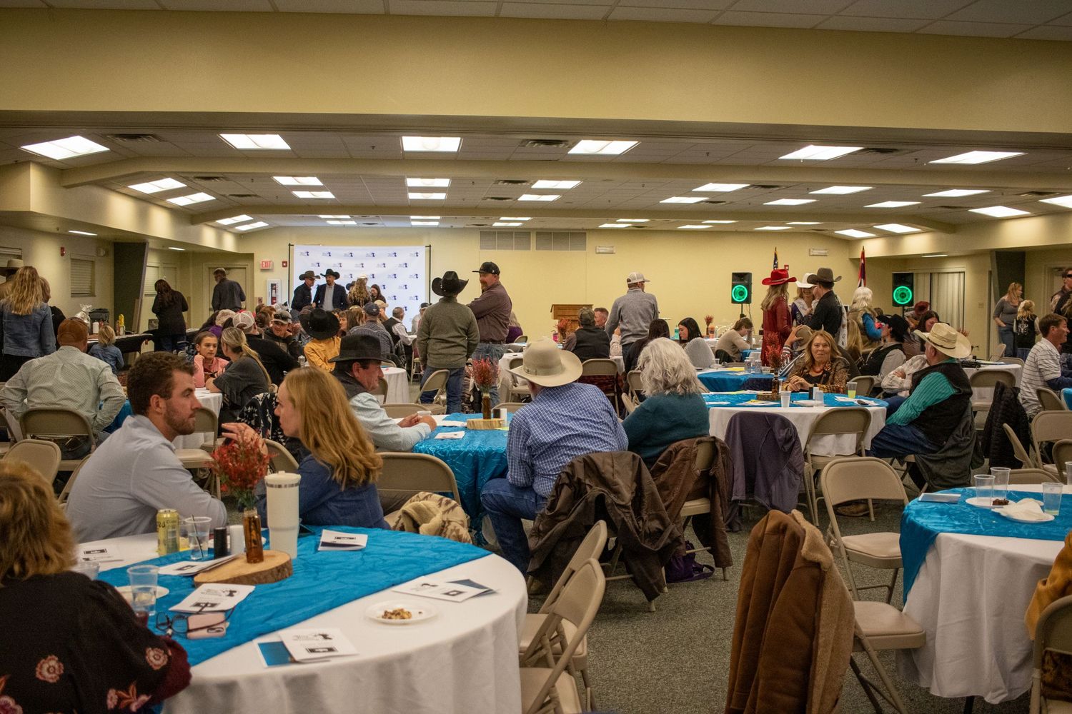 Large group gathered in a community room with round tables and blue tablecloths for a social event.