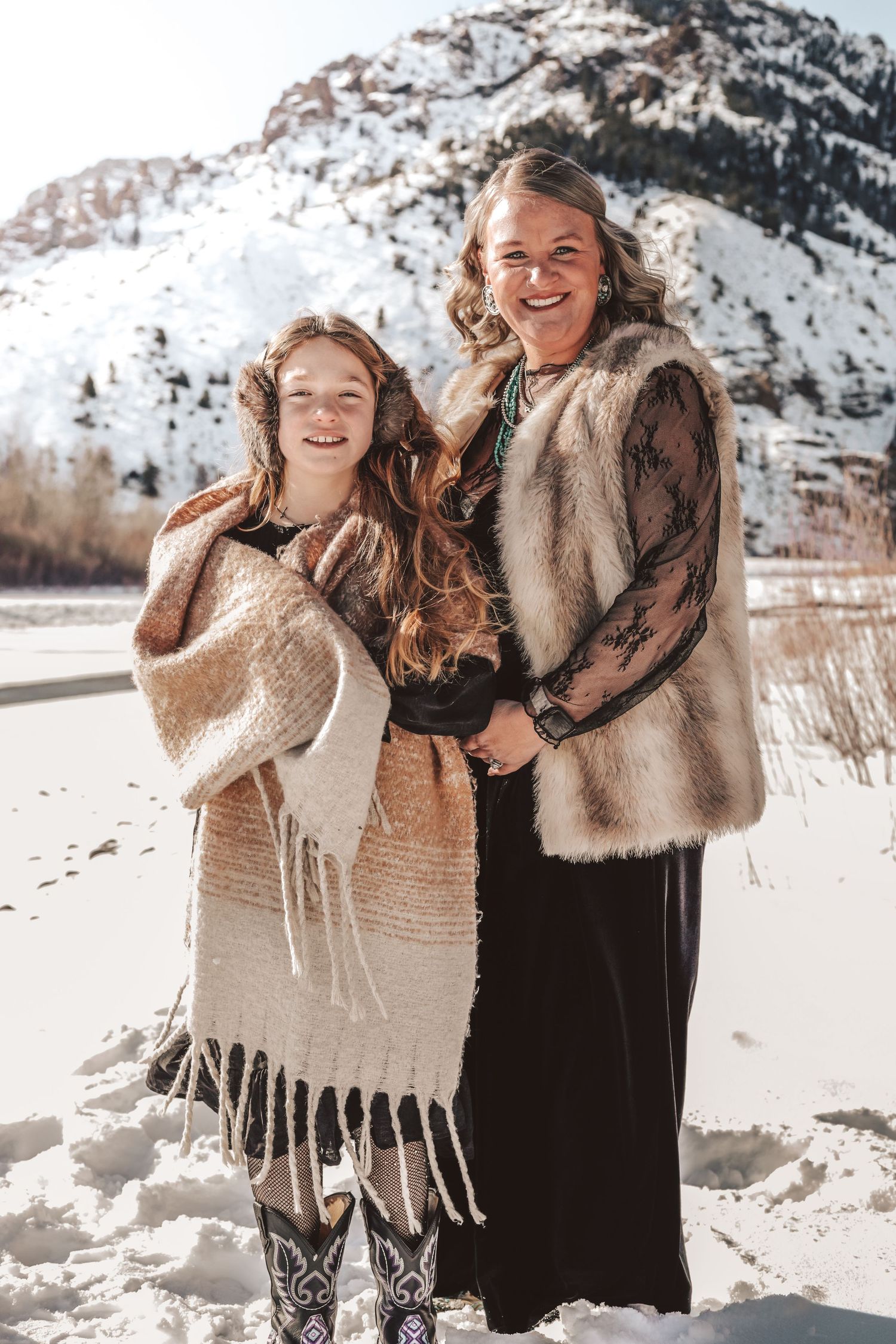 Two people in winter coats stand together smiling in front of a snow-covered mountain.