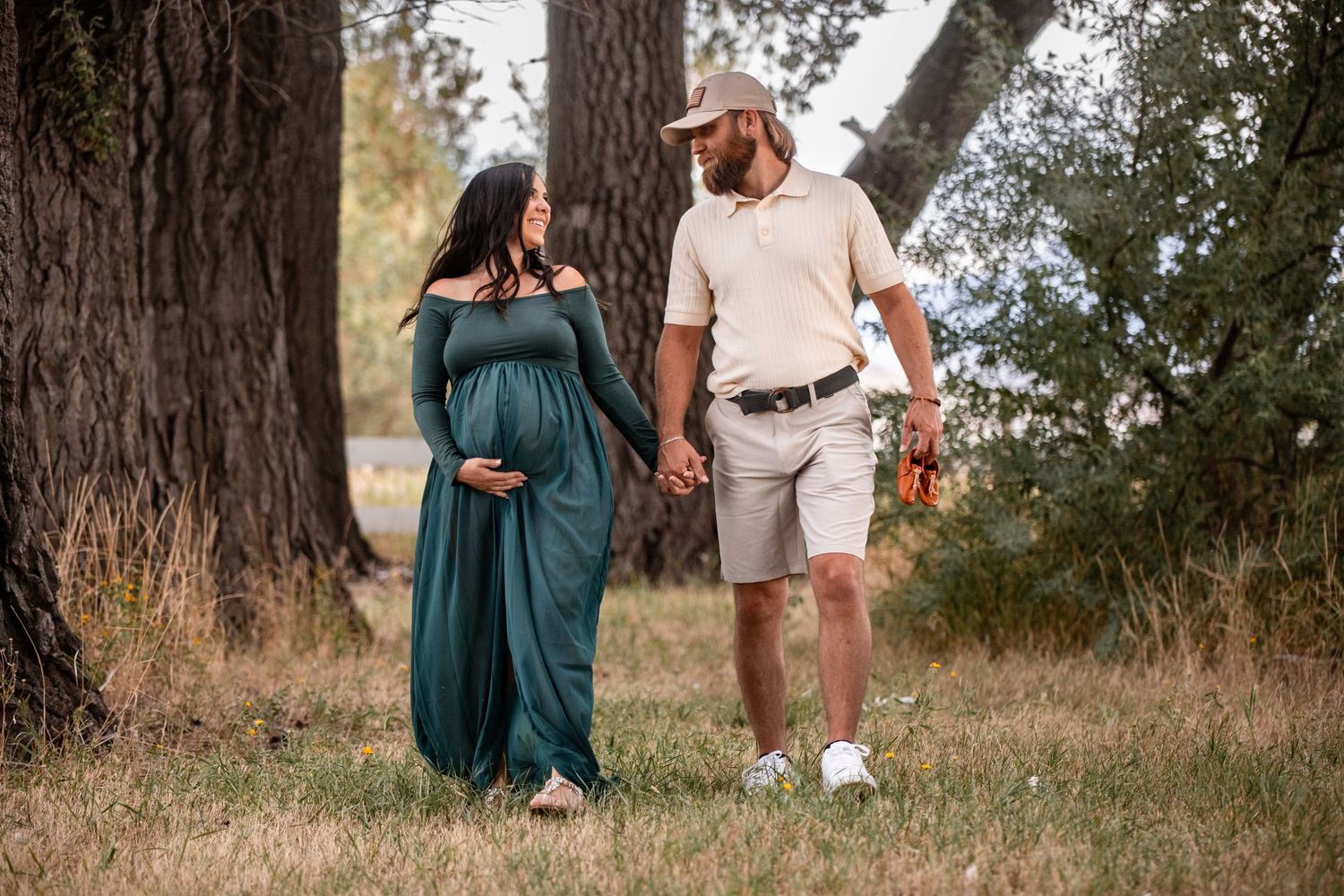 A couple stands together under tall trees in a natural outdoor setting during golden hour.