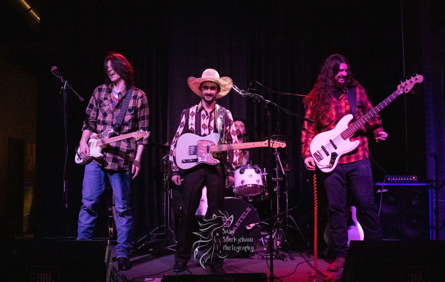 A country rock band performs on stage with guitars and drums under theatrical lighting.