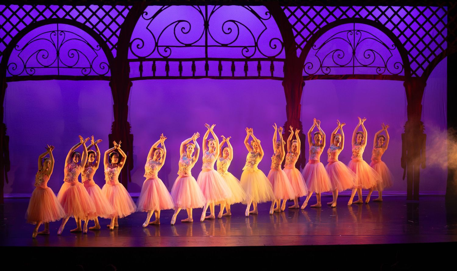 Line of ballerinas in white tutus perform under decorative arches with purple stage lighting.