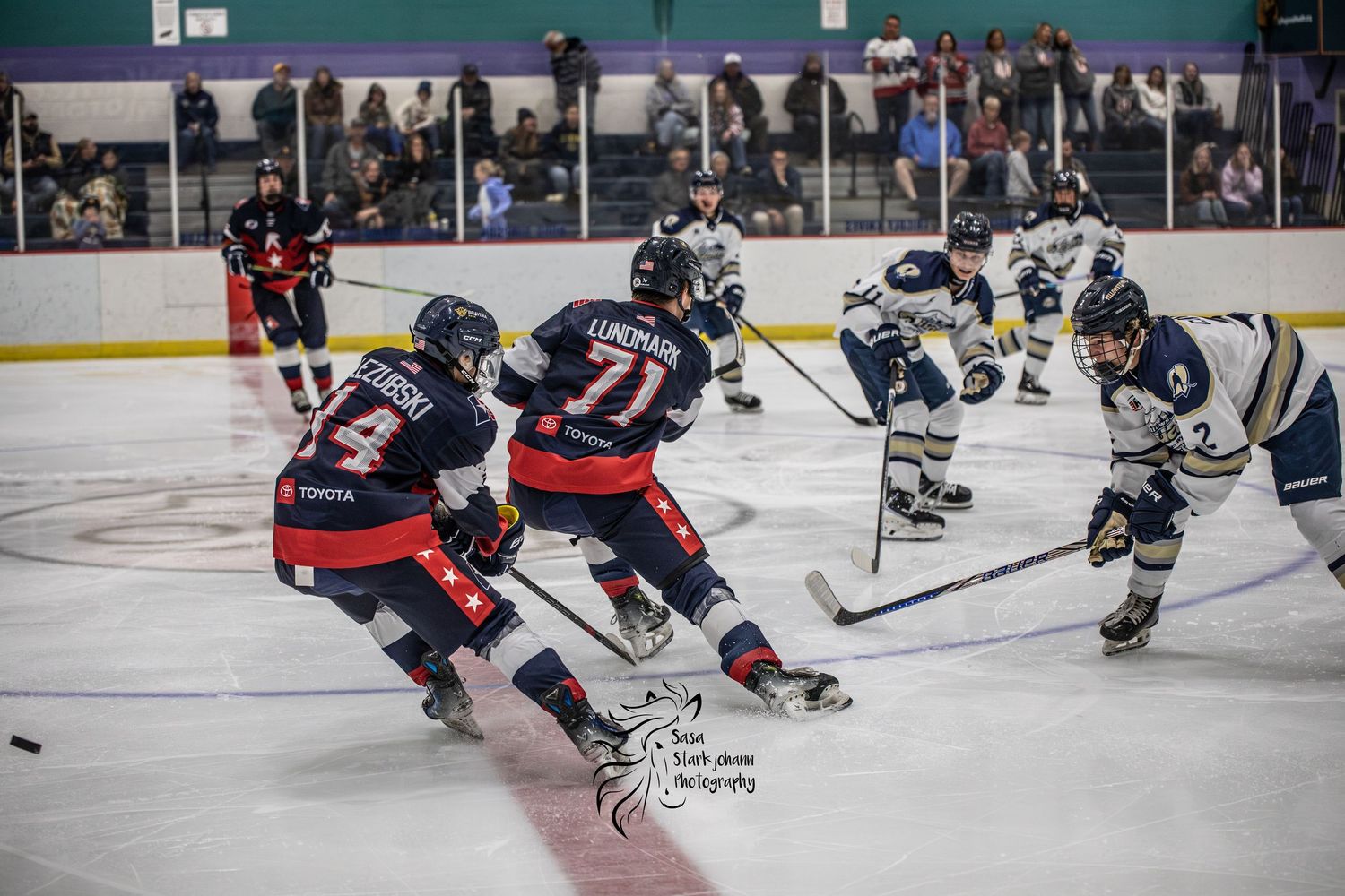 Youth ice hockey players compete on indoor rink, red team player #71 races toward goal against navy blue opponents.