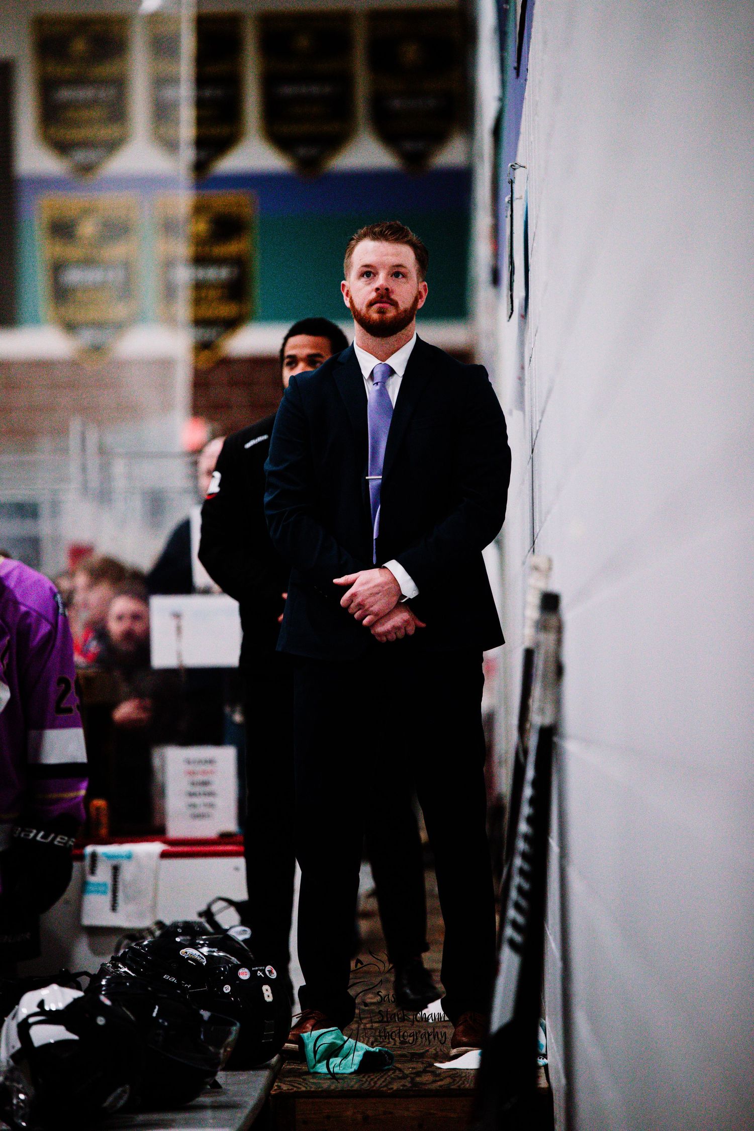 Coach in suit and tie observes from behind hockey bench during game.