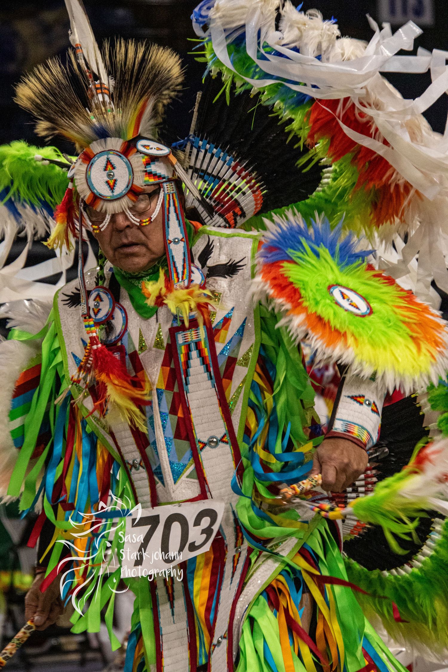 Powwow dancer in vibrant multicolored feathered regalia with competition number performing traditional dance.