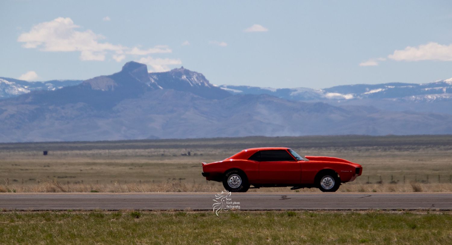 Red classic muscle car driving on empty road with mountains in background.