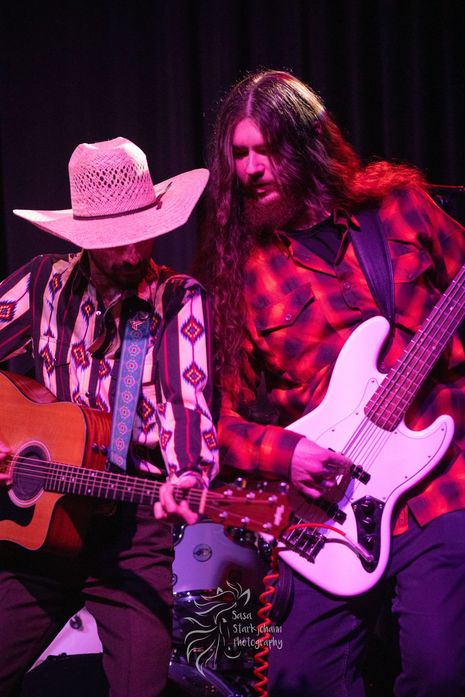 Musicians performing on stage with acoustic and electric guitars, one wearing a cowboy hat.
