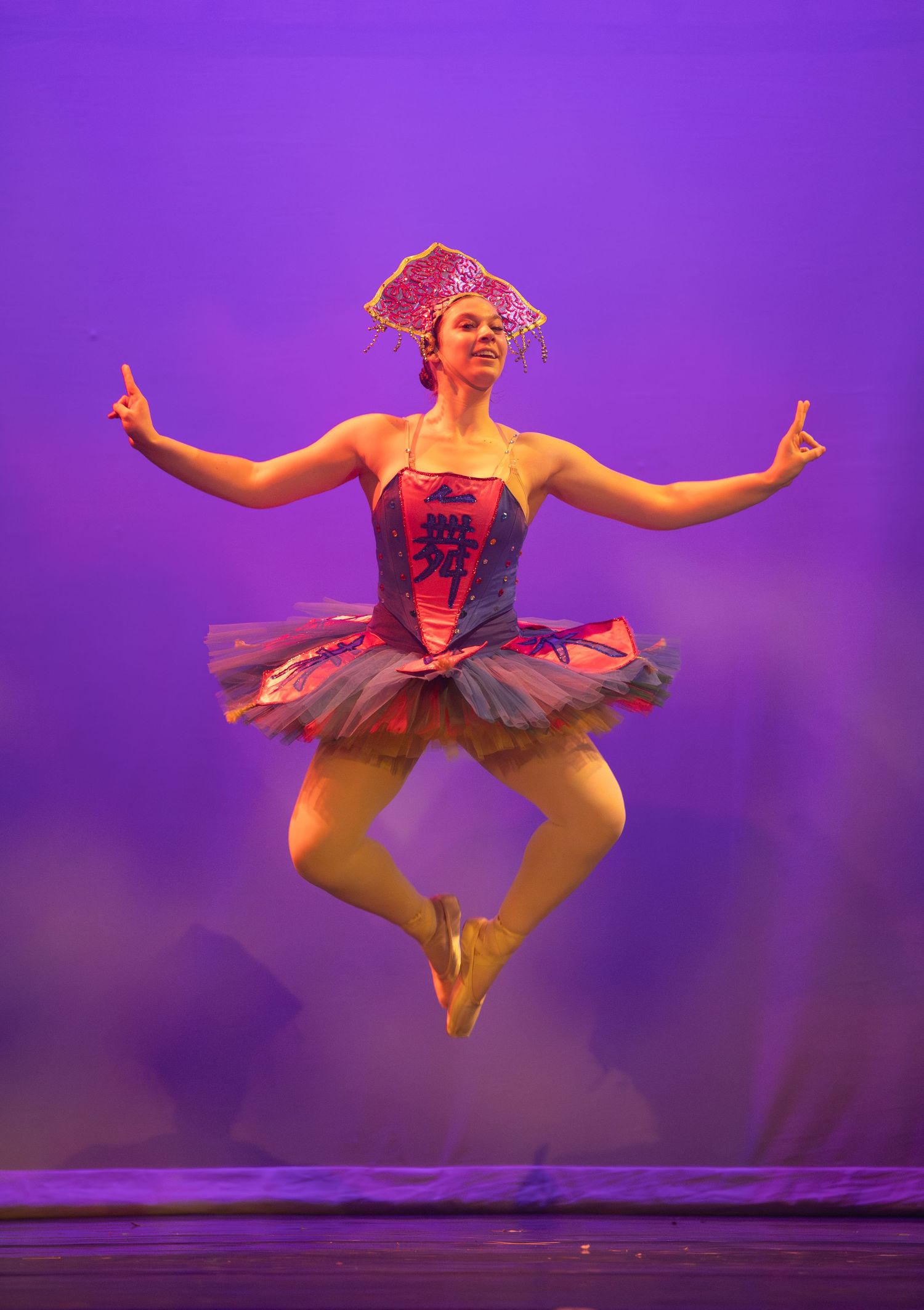 Ballet dancer in red tutu leaps gracefully against purple stage backdrop.