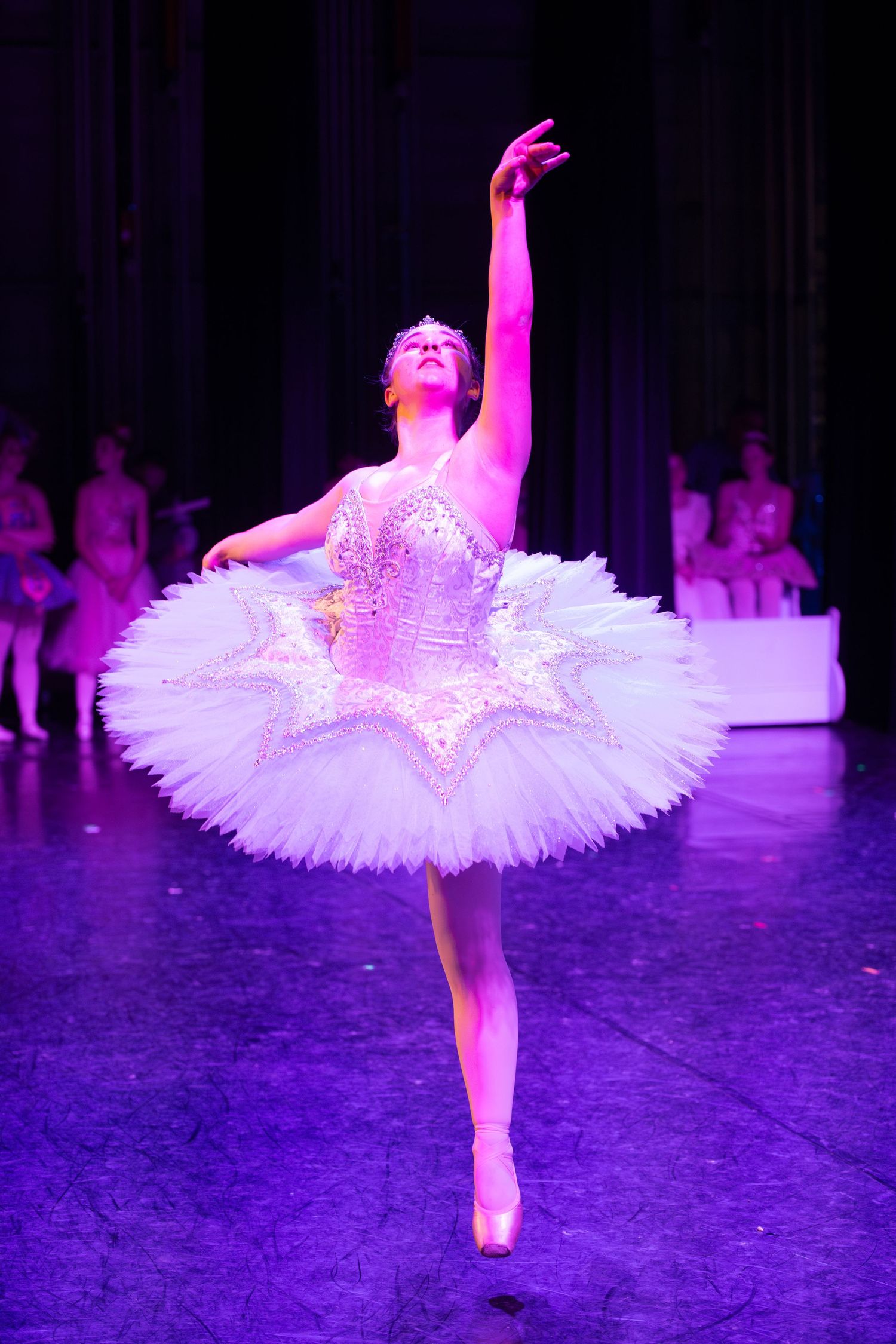 Ballet dancer in white tutu performs graceful pose under purple stage lighting.