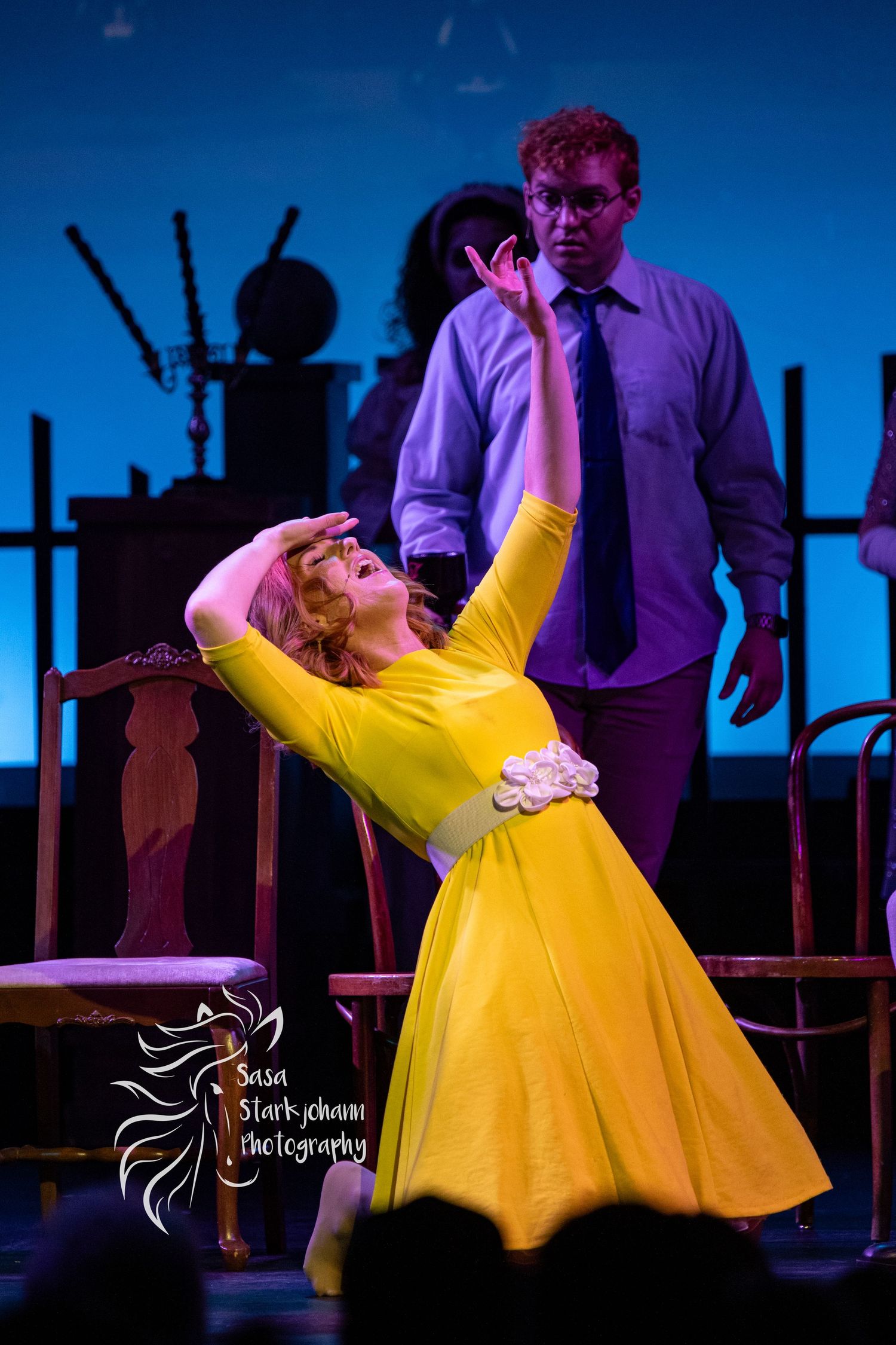 Theatrical performance with performers in yellow dress and purple shirt on stage with dramatic lighting.