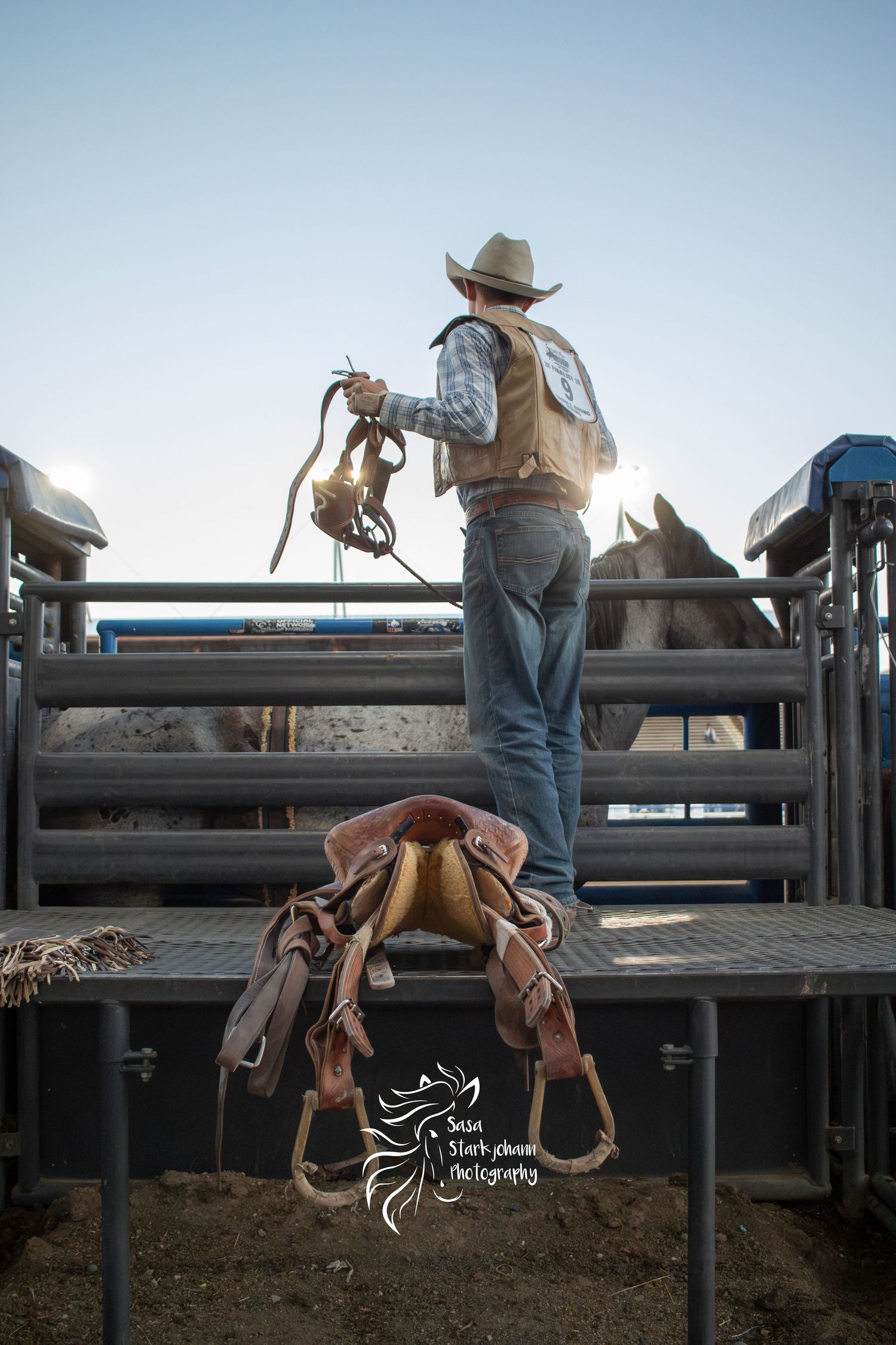 Cowboy in western gear inspecting rodeo equipment near horse stalls at sunset.