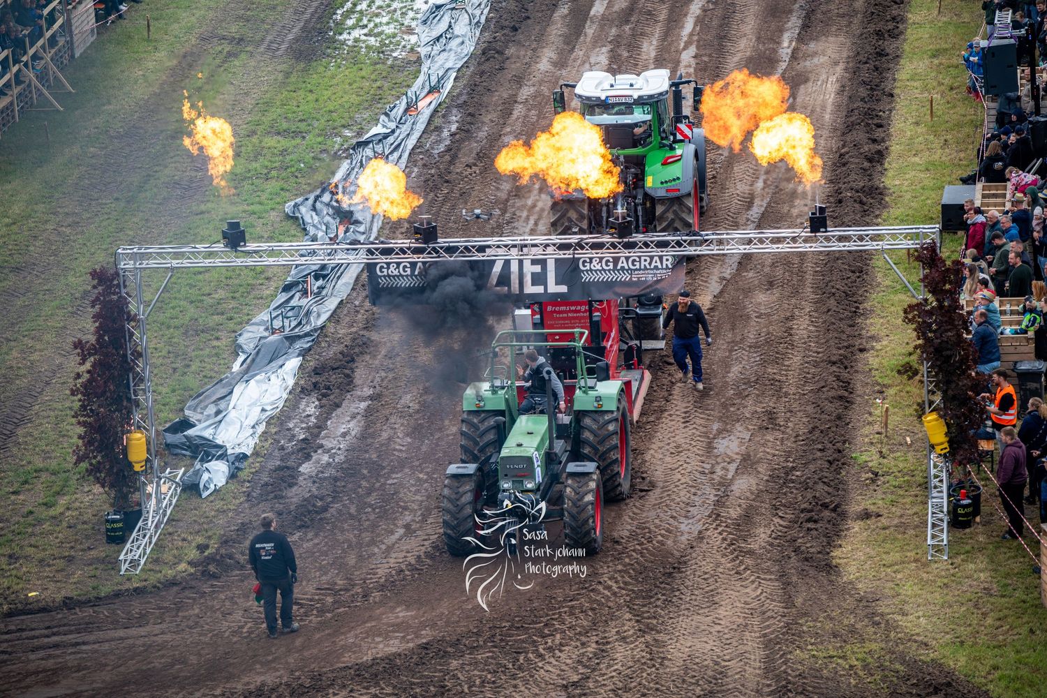 Two green tractors race on a dirt track with dramatic fire jets shooting from the sides at a tractor pulling competition.