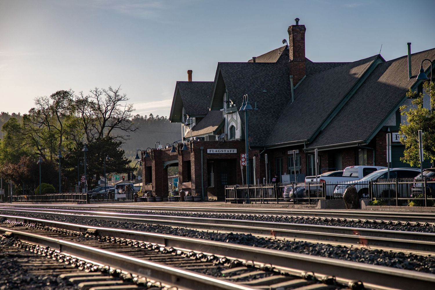 Historic train station with dark slate roof along railroad tracks at sunset with parked cars in the background.