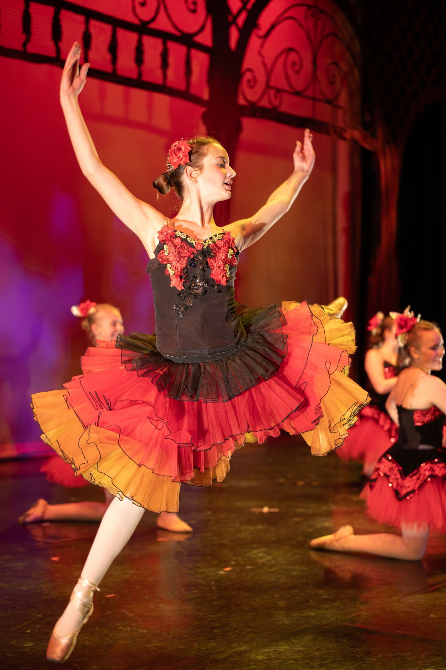 Ballet dancer performs elegant pose in red and black layered tutu under stage lighting.