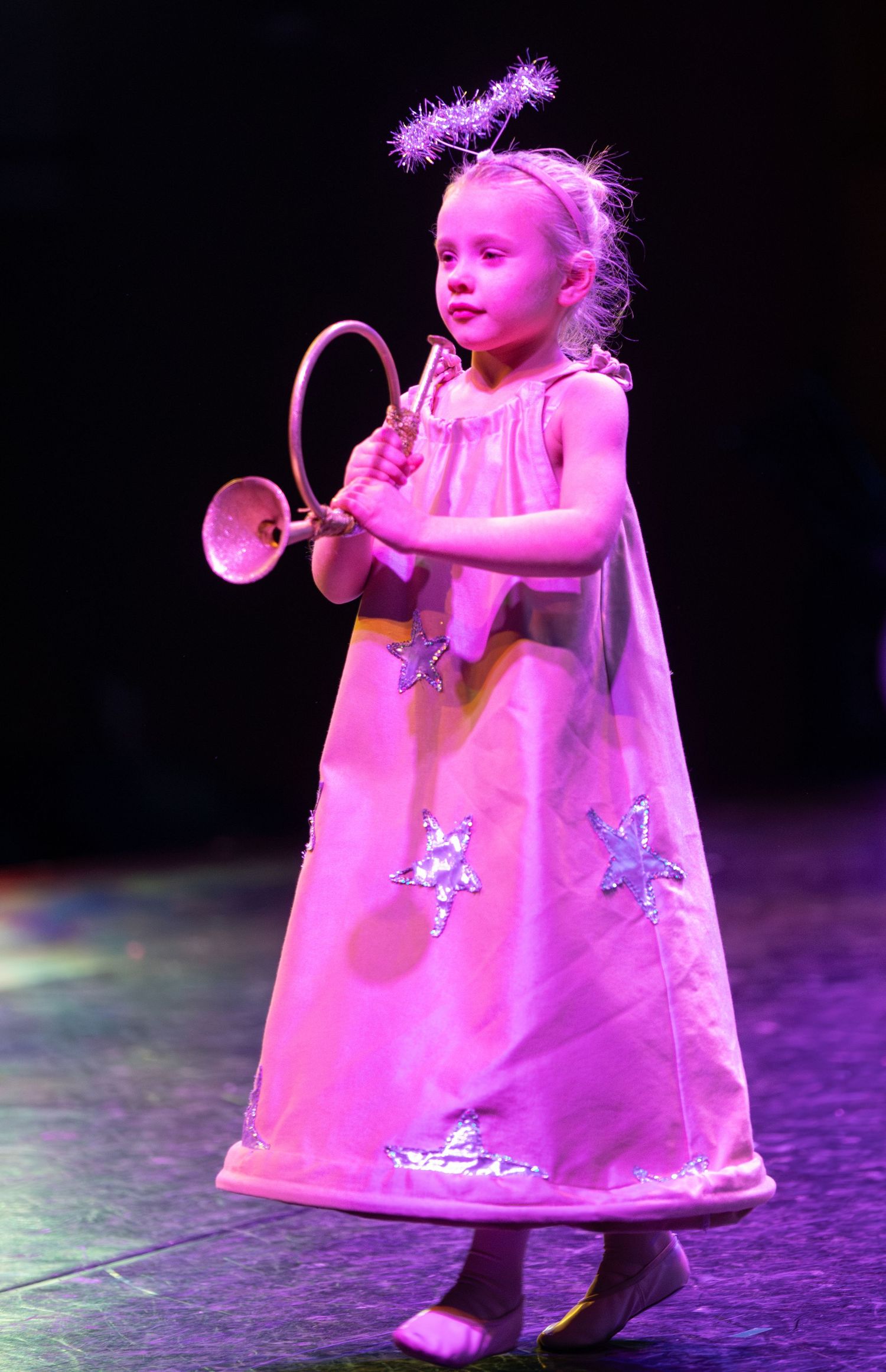 Young performer in pink dress with horn prop performs on stage under pink lighting.