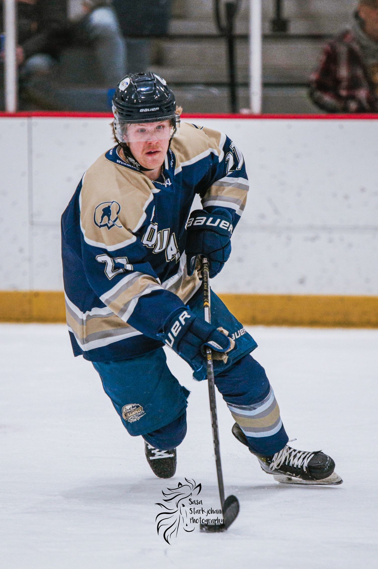 Hockey player in blue and tan jersey skating with puck on ice rink.
