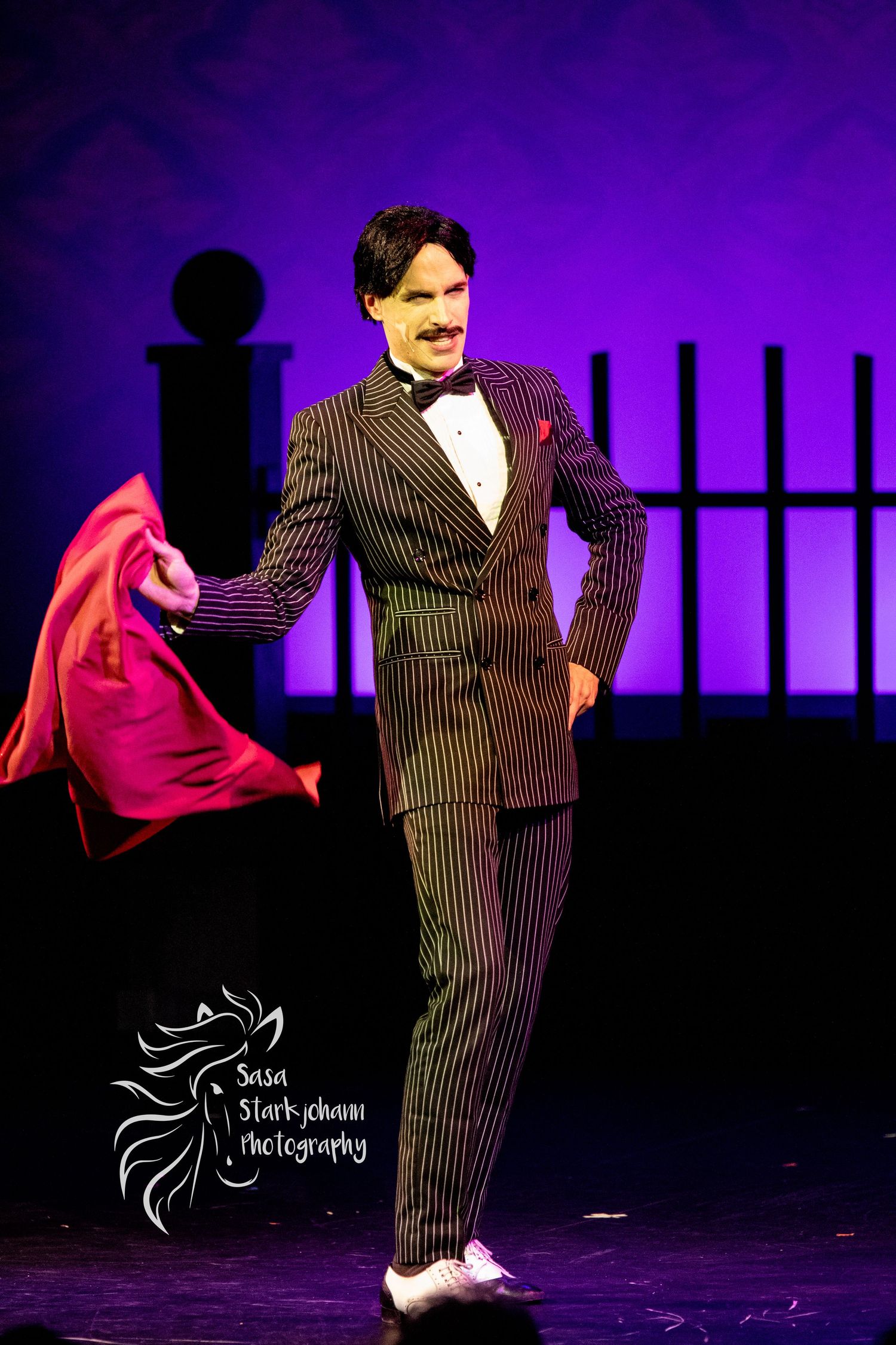 Stage performer in pinstriped suit gesturing dramatically against purple lighting.