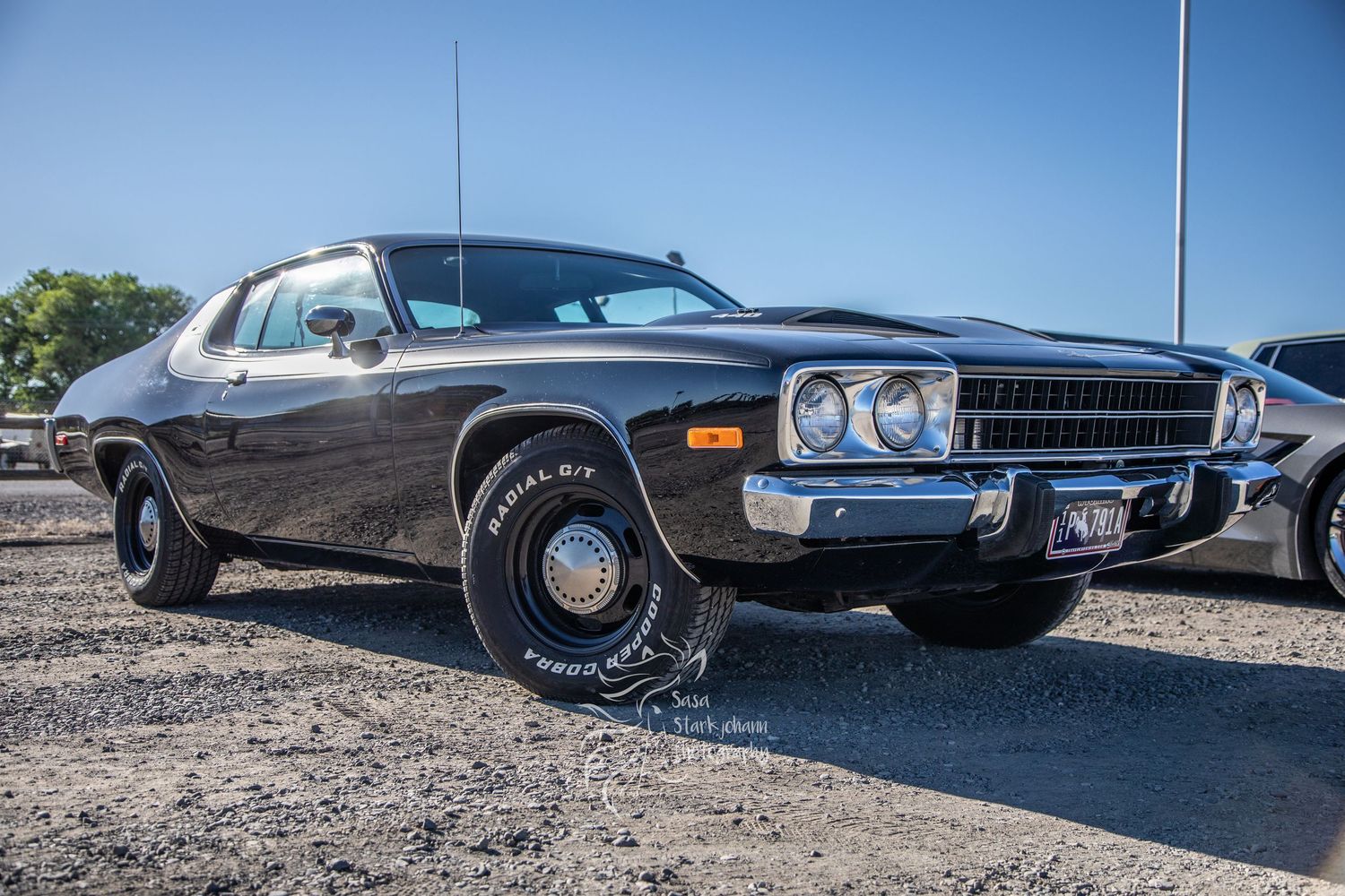 Classic brown Plymouth Road Runner muscle car with wide tires parked on gravel under blue sky.