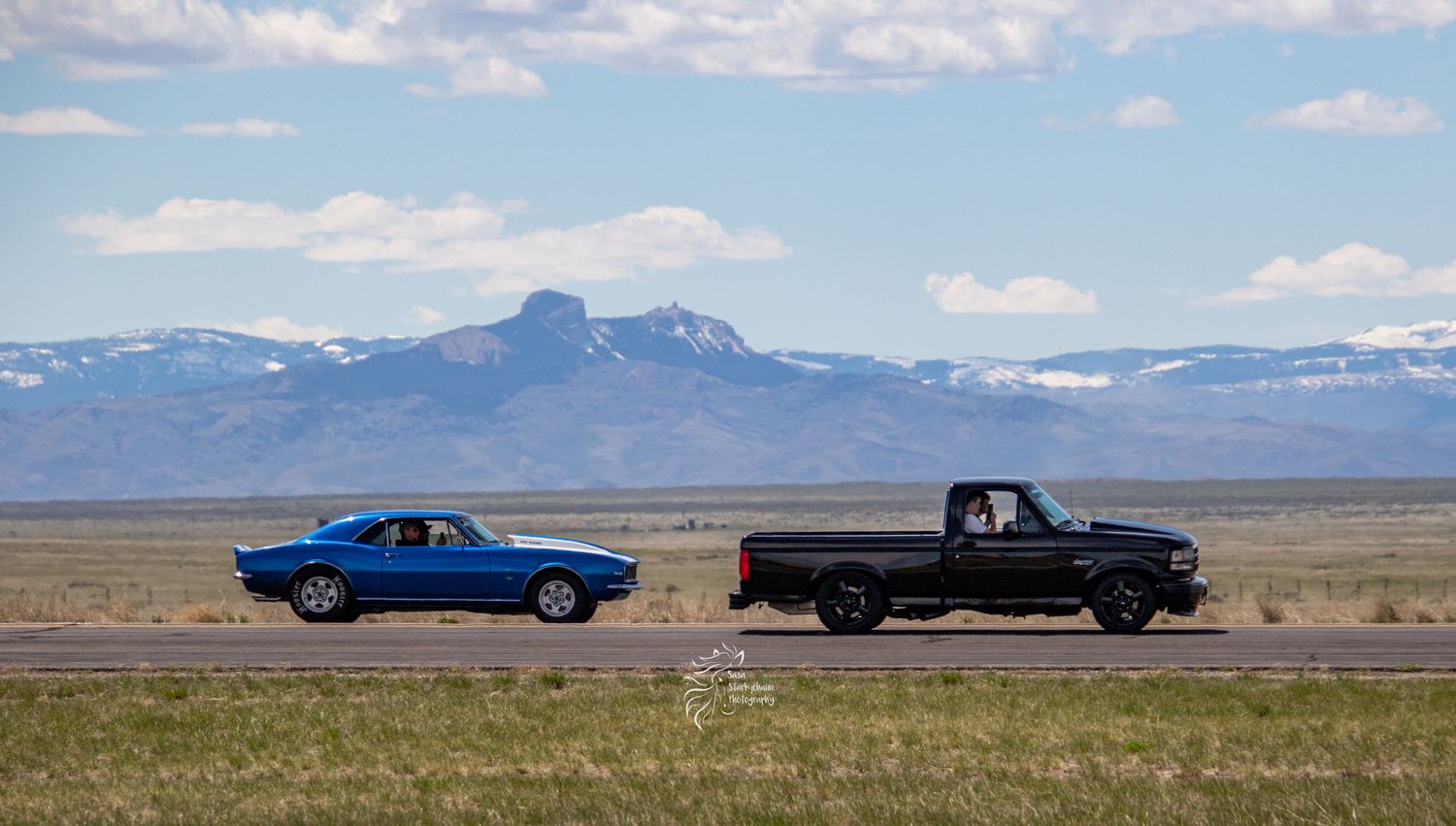 Blue classic car and black pickup truck driving on desert highway with mountains in background.
