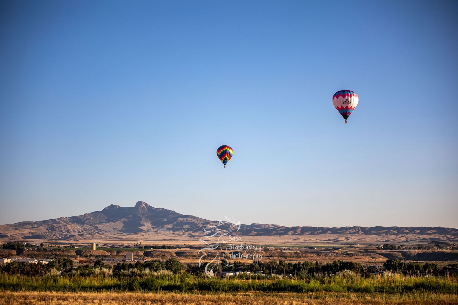 Two red hot air balloons float above a desert landscape with mountains and scrubland under clear blue skies.
