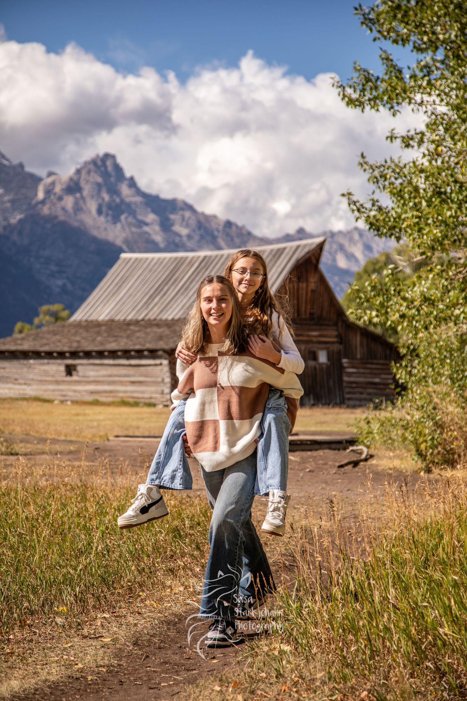 Two people enjoying a sunny day in front of a rustic barn with the Grand Teton mountains rising majestically in the background.