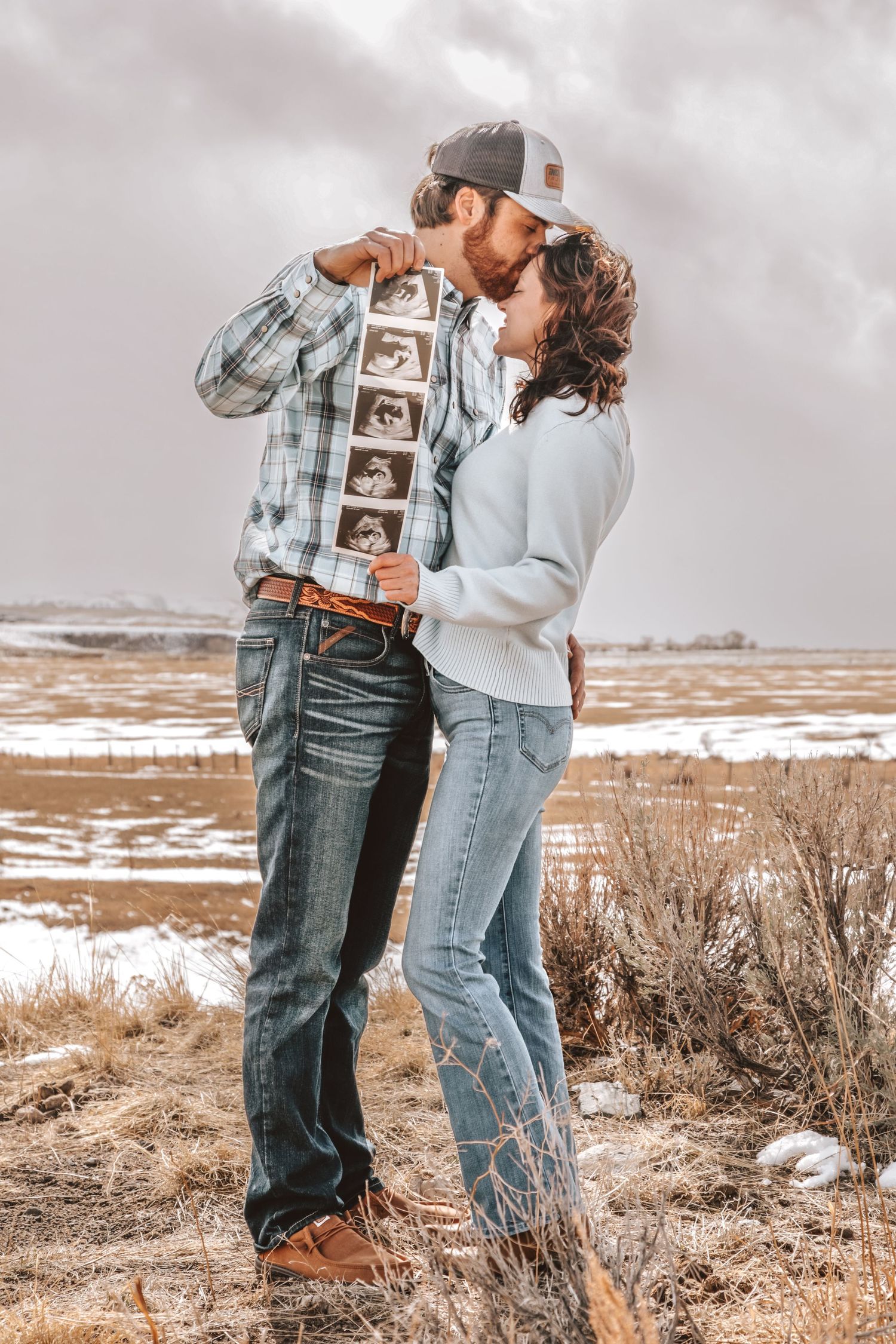 Couple embraces outdoors while holding ultrasound photos on a plaid shirt.