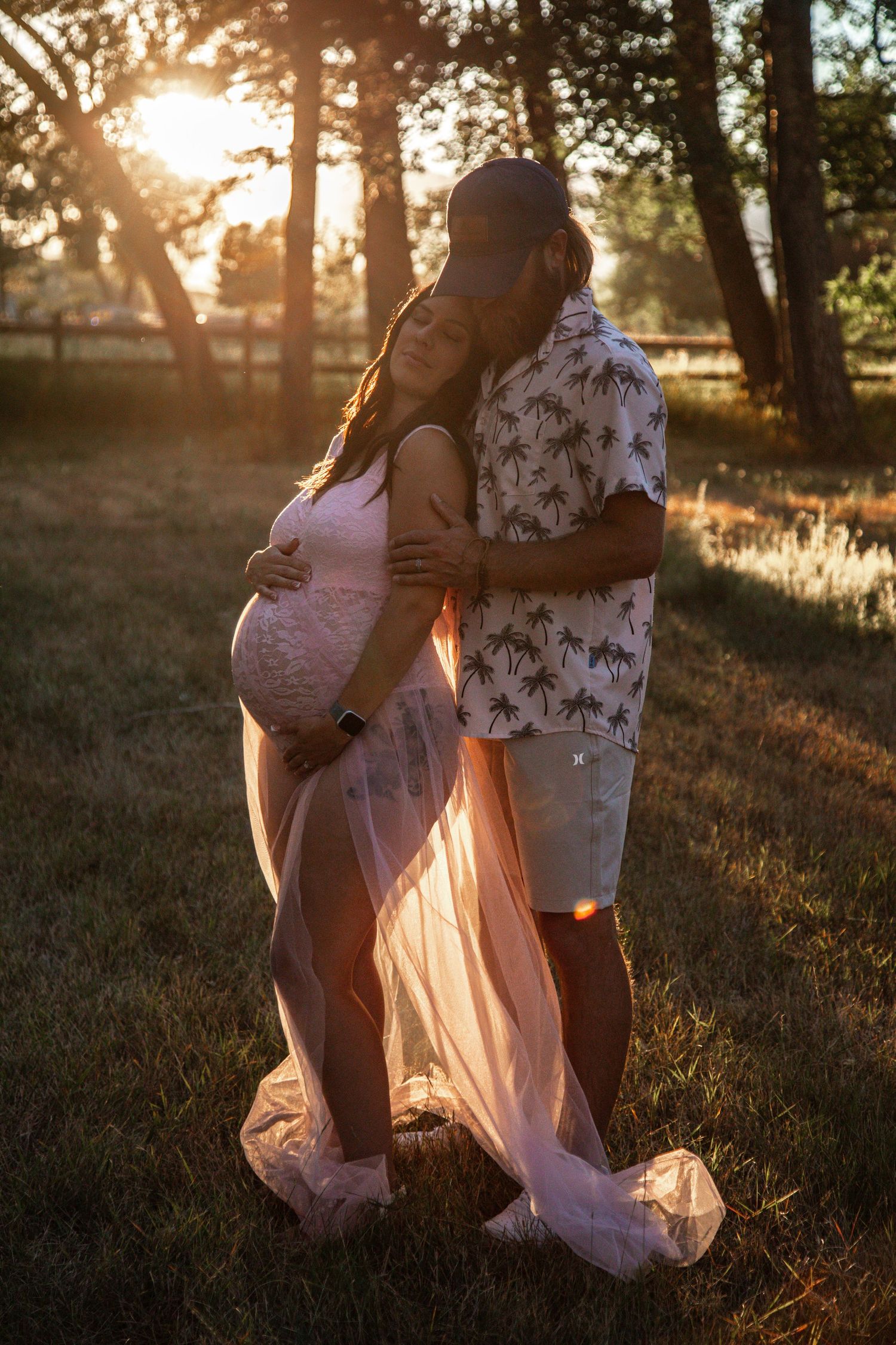 A couple in flowy summer outfits embraces during golden hour in a sunlit forest setting.